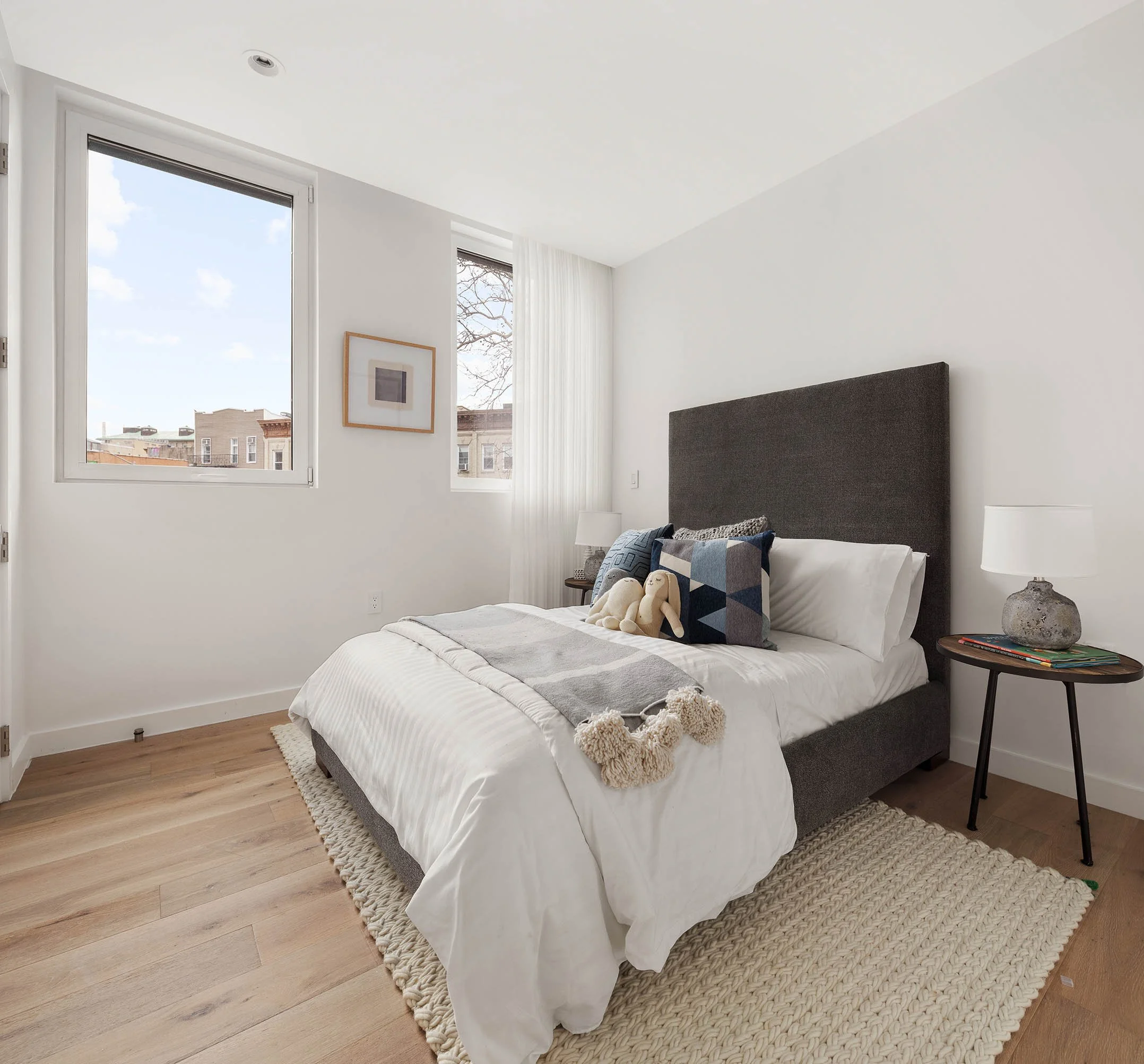 A bedroom with a large bed, dark gray headboard, white bedding, decorative pillows, a small stuffed animal, a gray throw with tassels, a cozy beige rug, a round side table with a lamp and books, large windows letting in natural light, and a framed pi