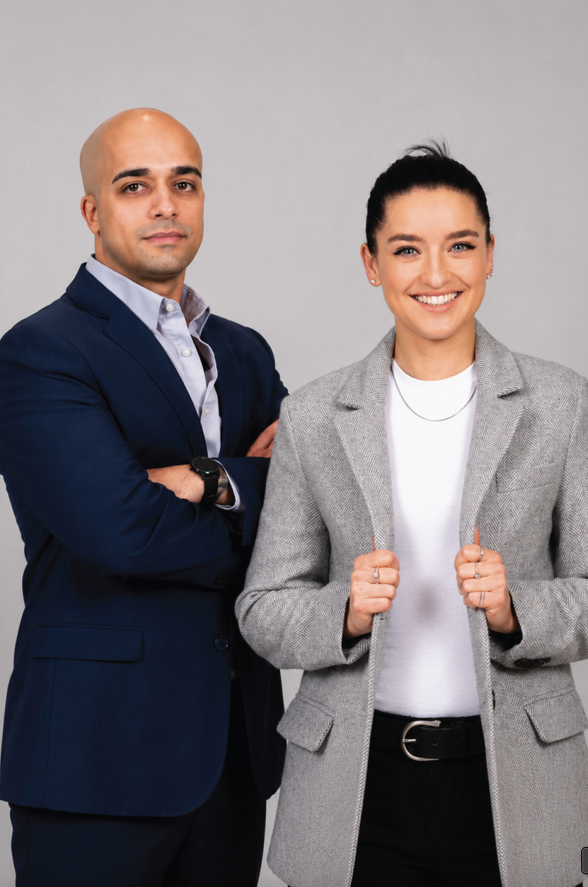 A confident woman smiling and wearing a gray blazer with a white shirt and black belt, standing with her fists raised. A serious man in a blue suit with a watch, arms crossed, stands next to her against a plain gray background.