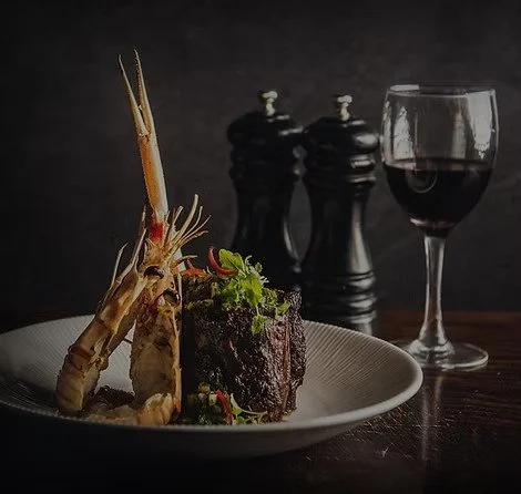 A plate with a cooked lobster tail, a grilled steak garnished with herbs, a glass of red wine, and black salt and pepper shakers on a dark background.