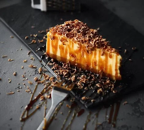 A slice of cheesecake topped with chocolate shavings and caramel drizzle on a black slate serving board, with chocolate shavings and drizzle scattered around.