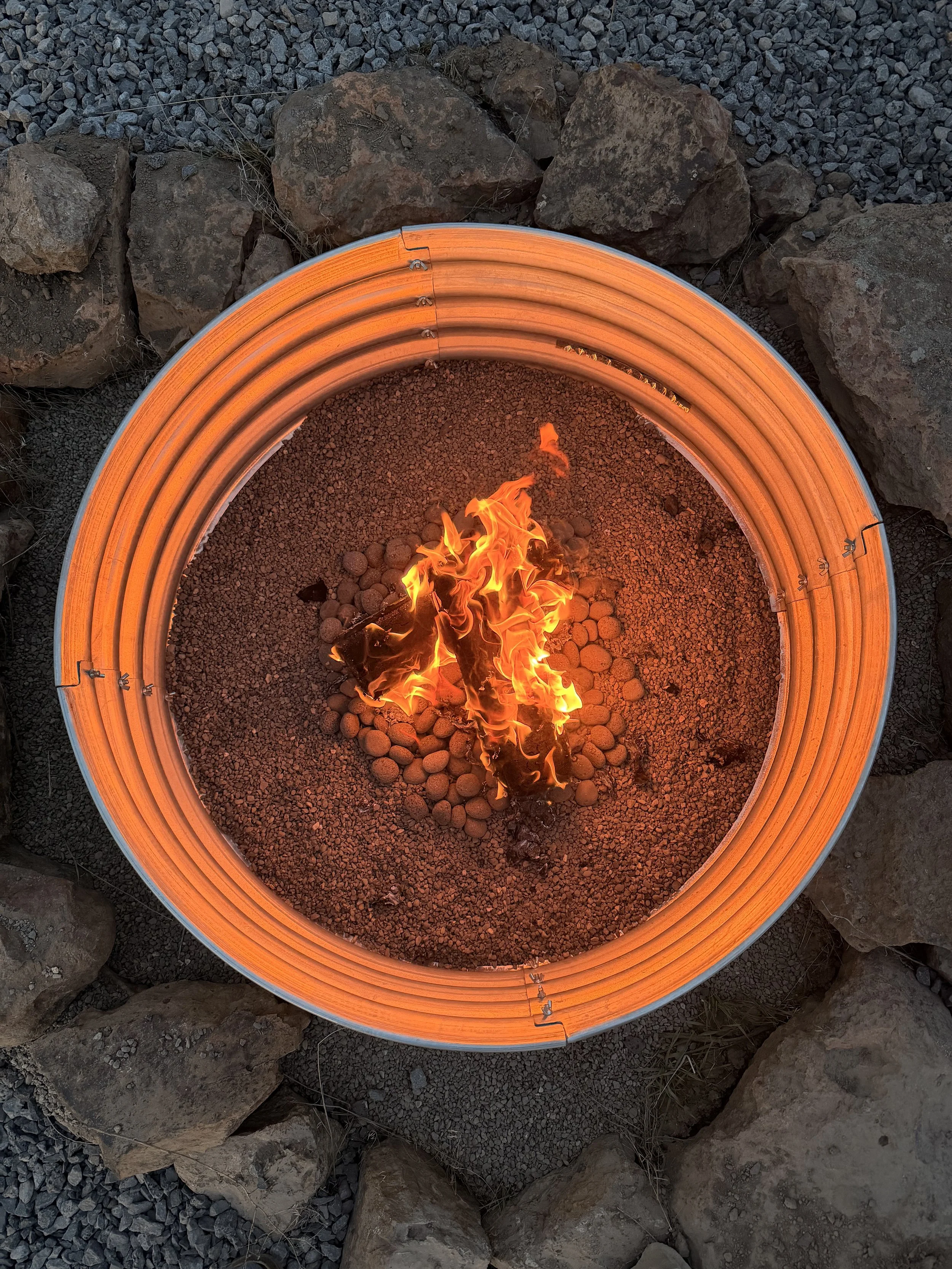 A circular fire pit with a glowing orange glow, surrounded by large rocks and smaller gravel at dusk.