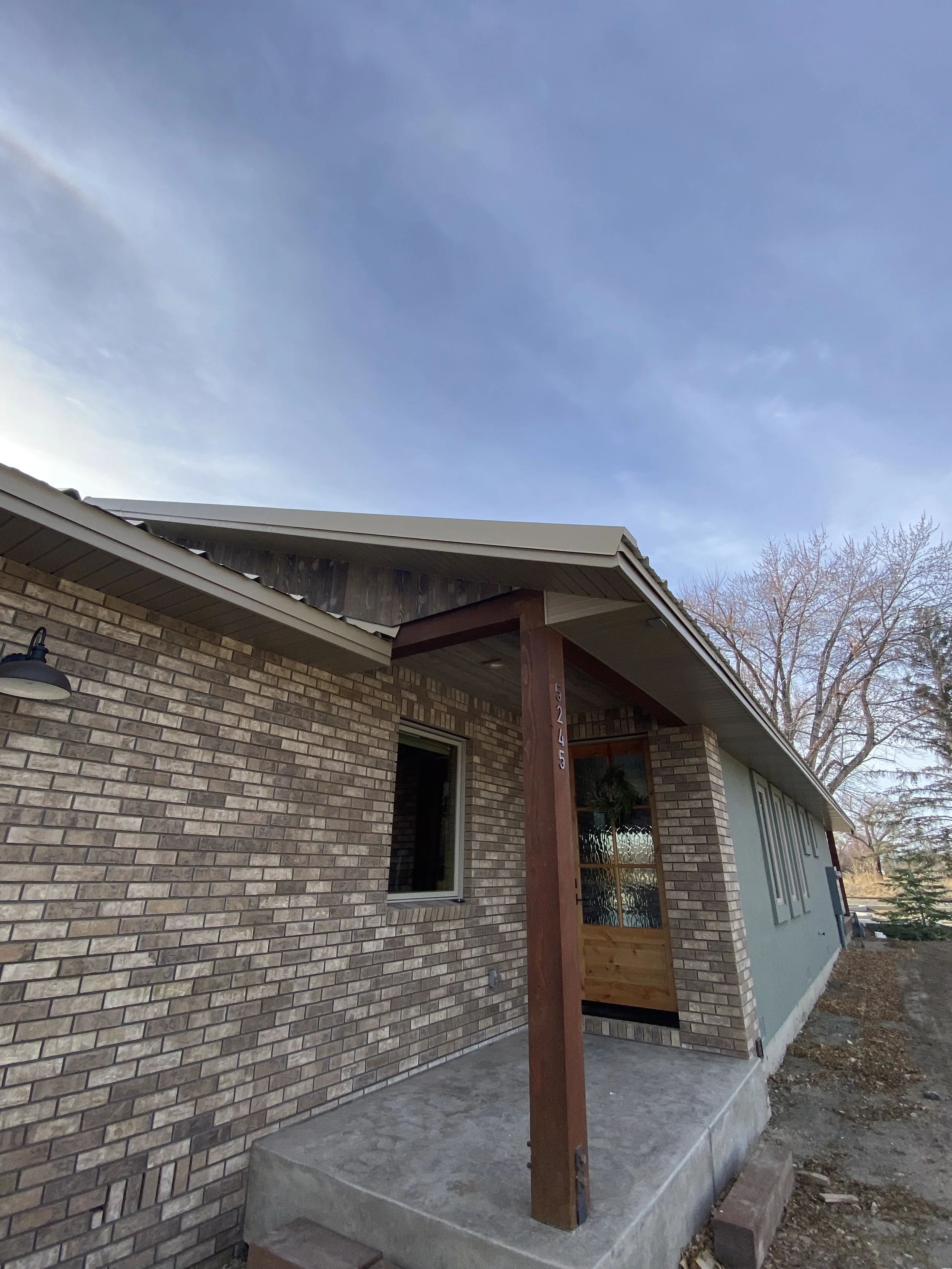 View of a house with a brick exterior, an elevated front porch with a concrete slab foundation, and a wooden post supporting the roof. The sky is mostly clear with some clouds, and leafless trees are visible in the background.