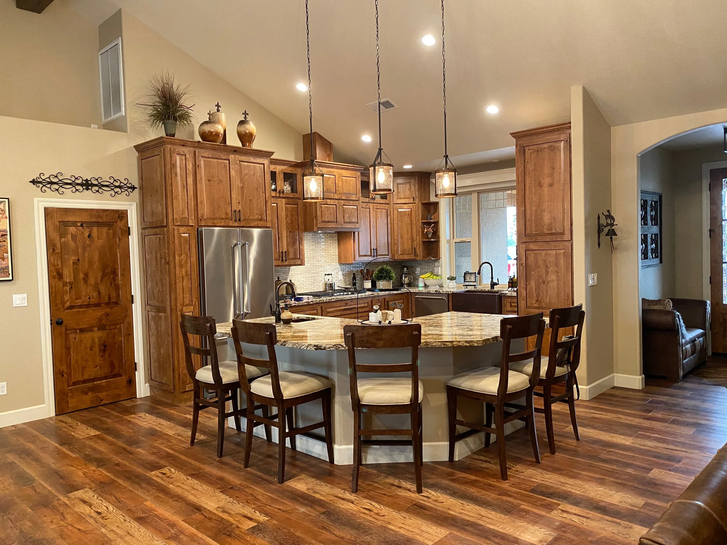 A kitchen with wooden cabinets, stainless steel refrigerator, and a round granite island with six chairs. There are three pendant lights hanging above the island and decorative vases on top of the cabinets. Recessed ceiling lights illuminate the space.