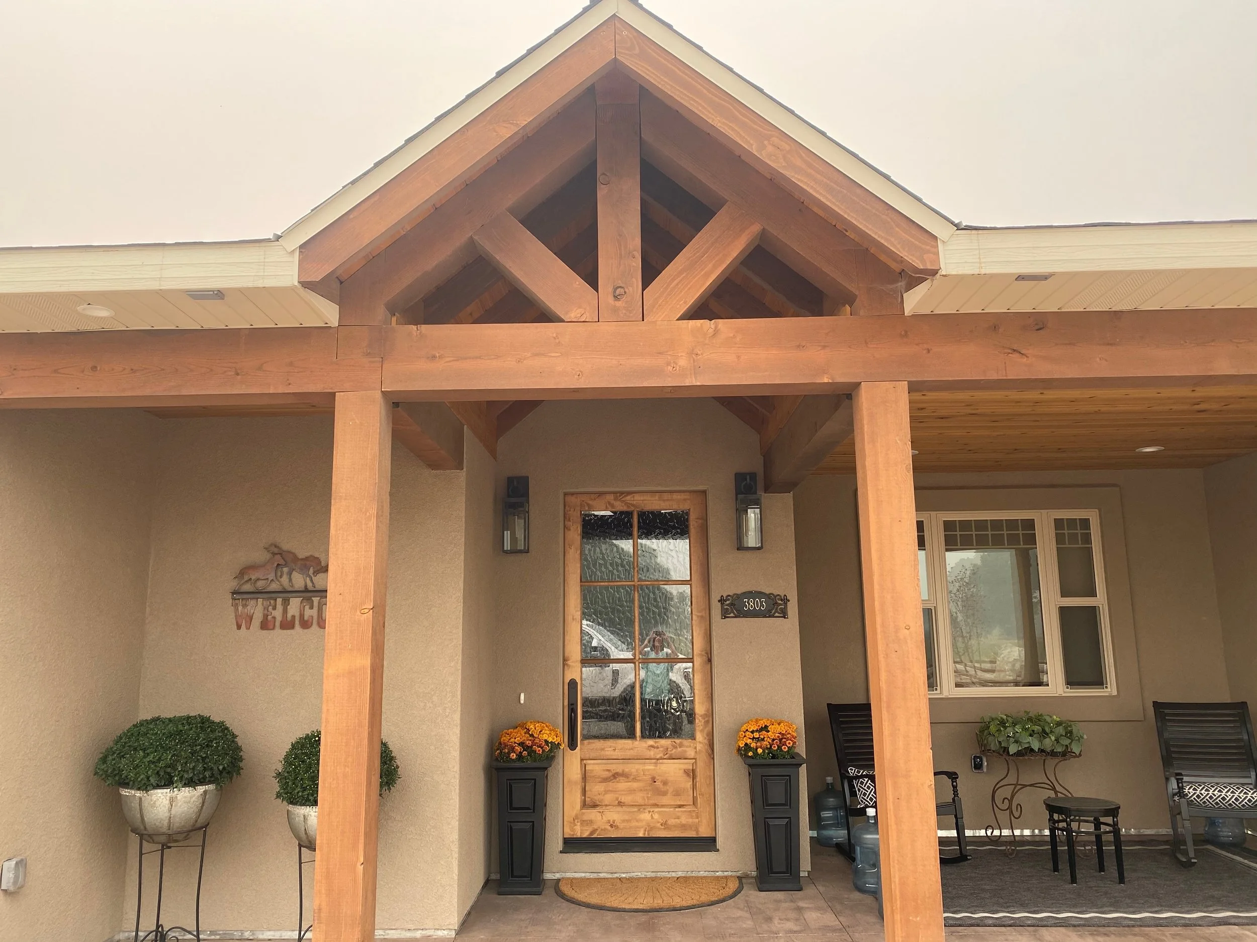 Close-up front porch view of a house entrance with a wooden door, potted plants, and outdoor furniture, under construction or renovation with exposed wood beams.