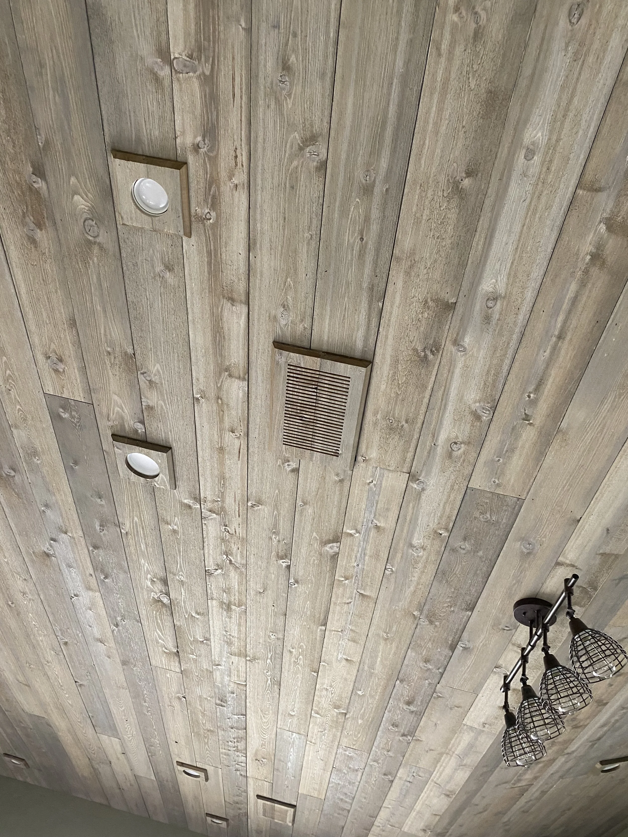 Ceiling made of wooden planks with lighting fixtures, ventilation grate, and a spotlight.