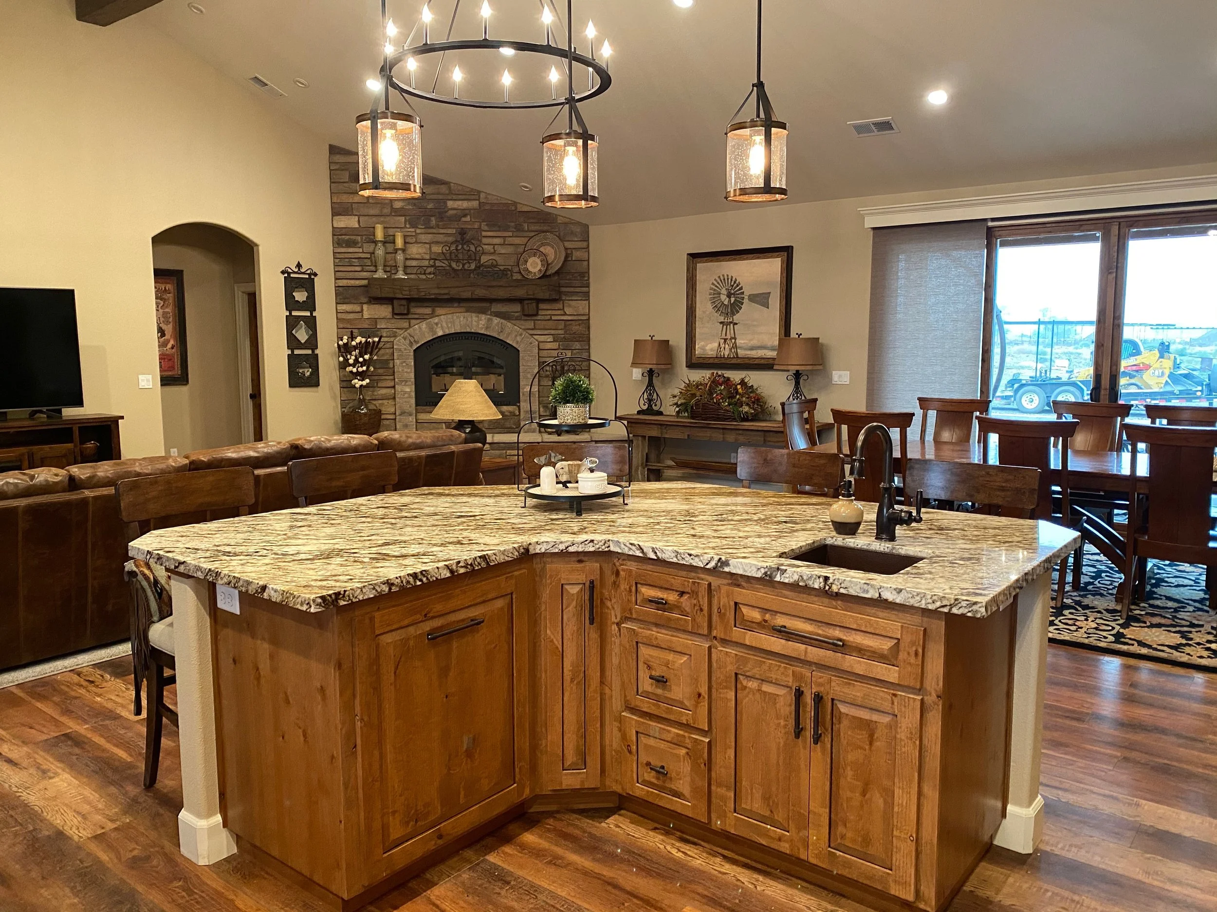 A spacious open-plan living and dining area with a kitchen island in the foreground, featuring a granite countertop and wooden cabinetry. The background includes a stone fireplace with decorative items on the mantle, framed artwork, and large windows letting in natural light. There are various lamps and decor pieces throughout, creating a cozy, rustic atmosphere.