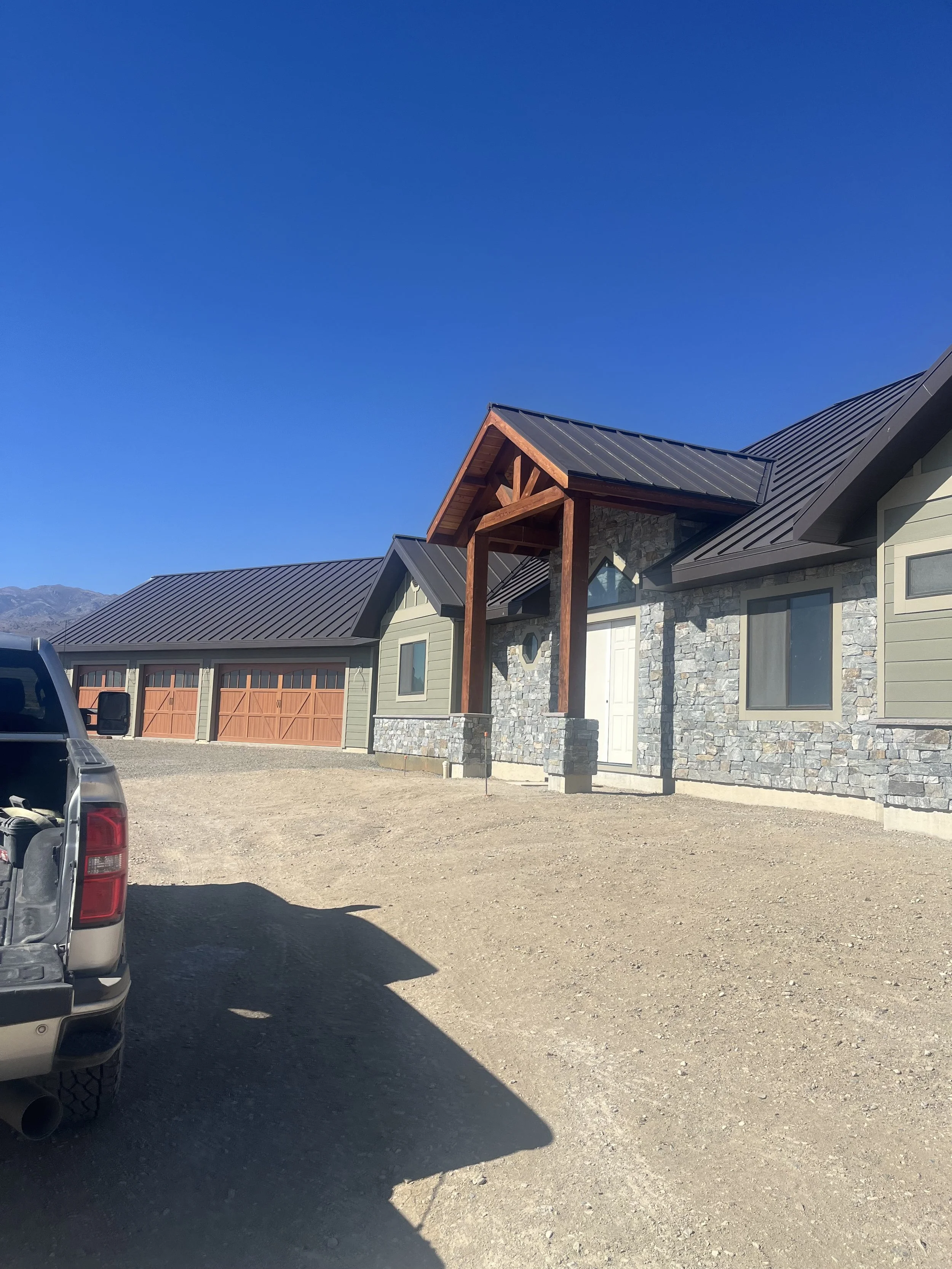 A house with a stone exterior and a metal roof, featuring a wooden entrance with a peaked roof, surrounded by an open dirt area with a vehicle partially visible on the left, under a clear blue sky.