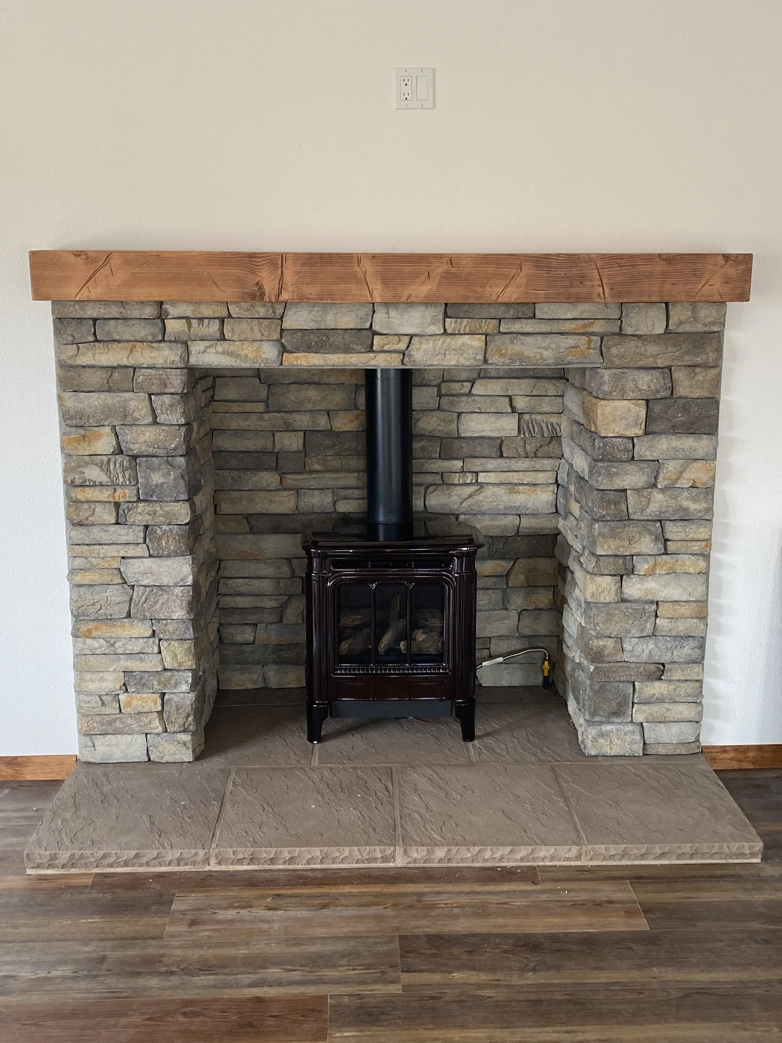 A stone fireplace with a wooden mantle, a black stove pipe, and a black stove. The fireplace is built with stacked stones and the mantle is made of a single wooden beam. The wall above the fireplace is plain and painted in a light color, with a power outlet and switch.