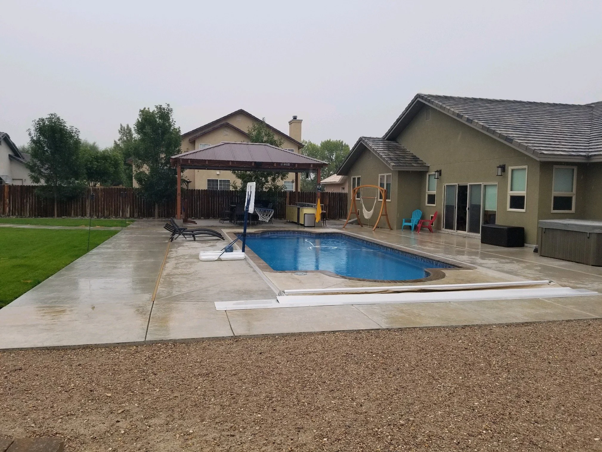 A backyard with a swimming pool, patio, and outdoor furniture on a cloudy, rainy day.