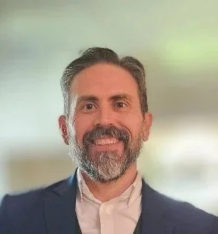 A middle-aged man with a beard and gray hair, wearing a dark blazer and white shirt, smiling in front of a light green background.