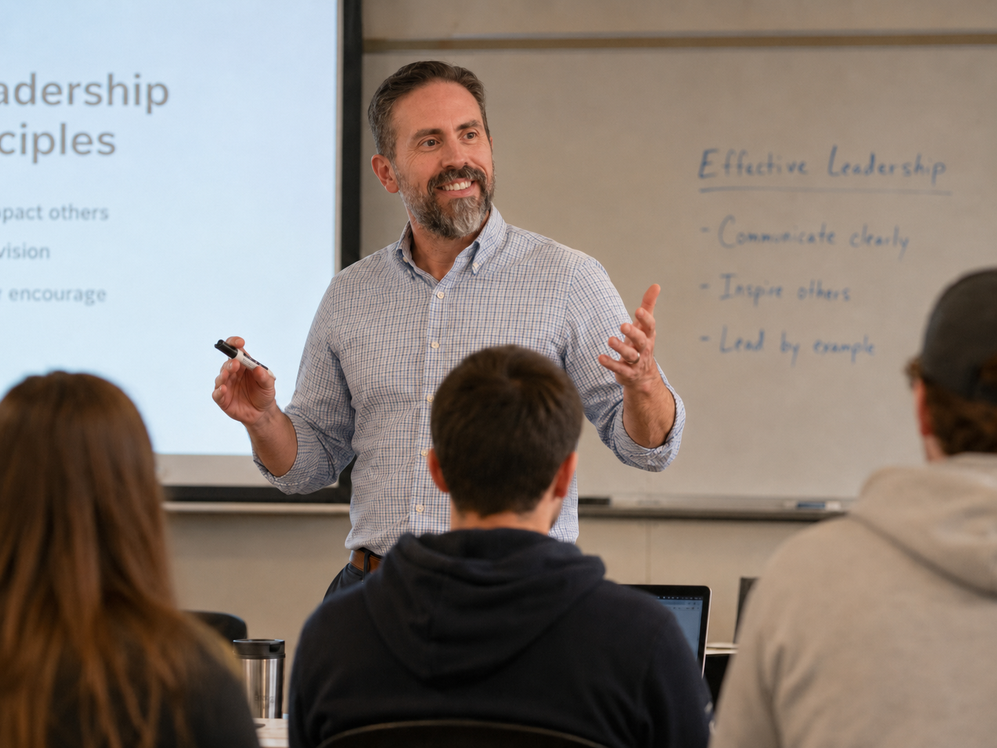 A man with a beard, wearing a checkered shirt, is giving a presentation in a classroom. He is holding a marker and smiling. Behind him is a whiteboard with handwritten notes about effective leadership and a projection screen displaying part of a presentation slide.