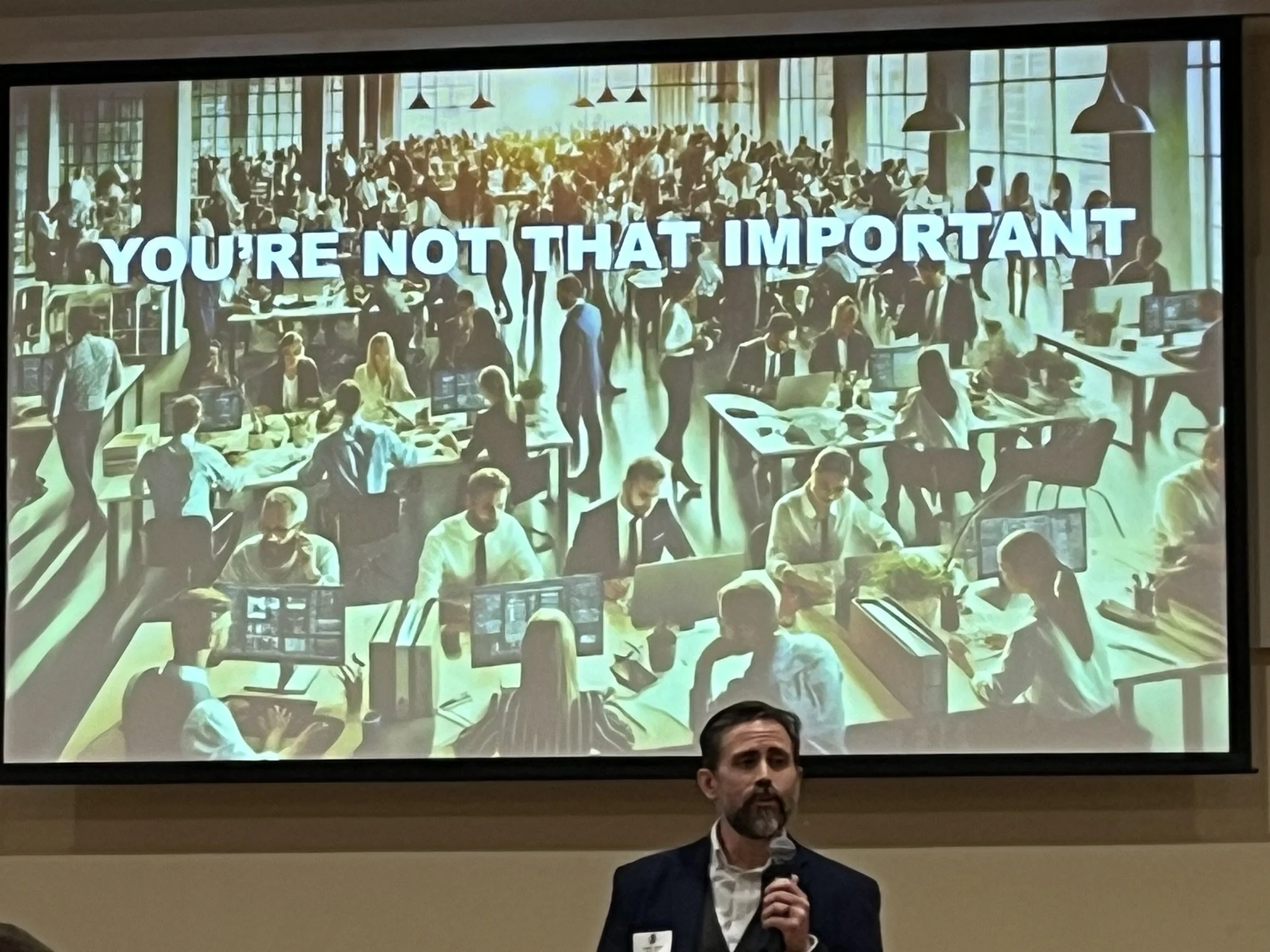A man with a beard and mustache holding a microphone and standing in front of a large screen showing a crowded office environment with many people working at desks.