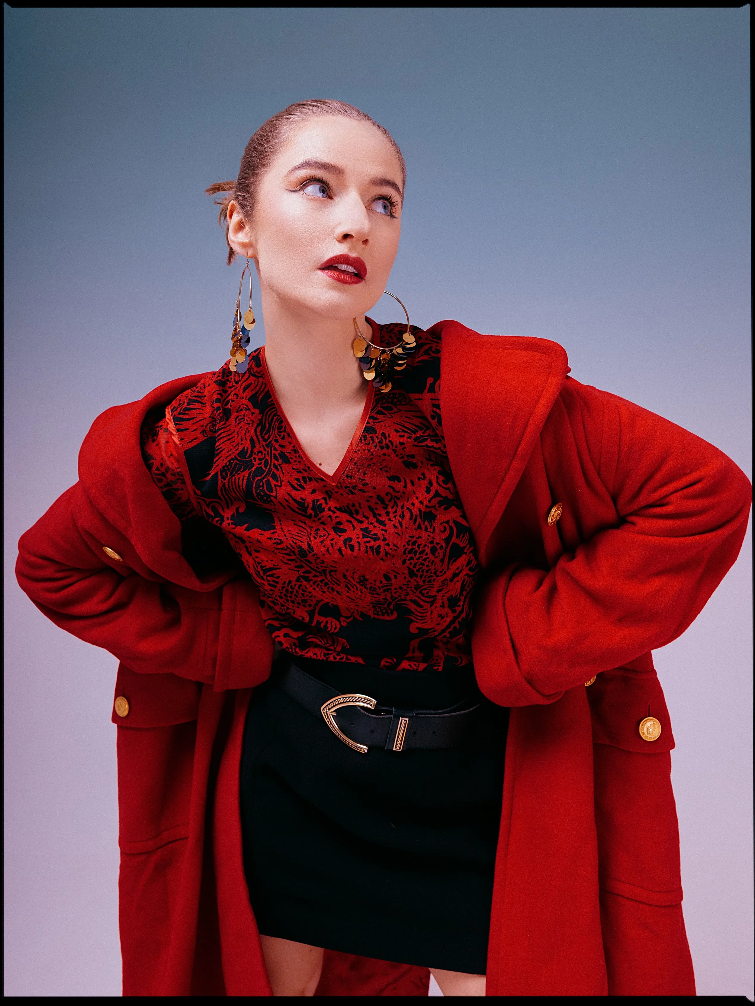 A woman dressed in a red coat and black dress with a leopard print pattern, accessorized with large hoop earrings, standing against a plain background.