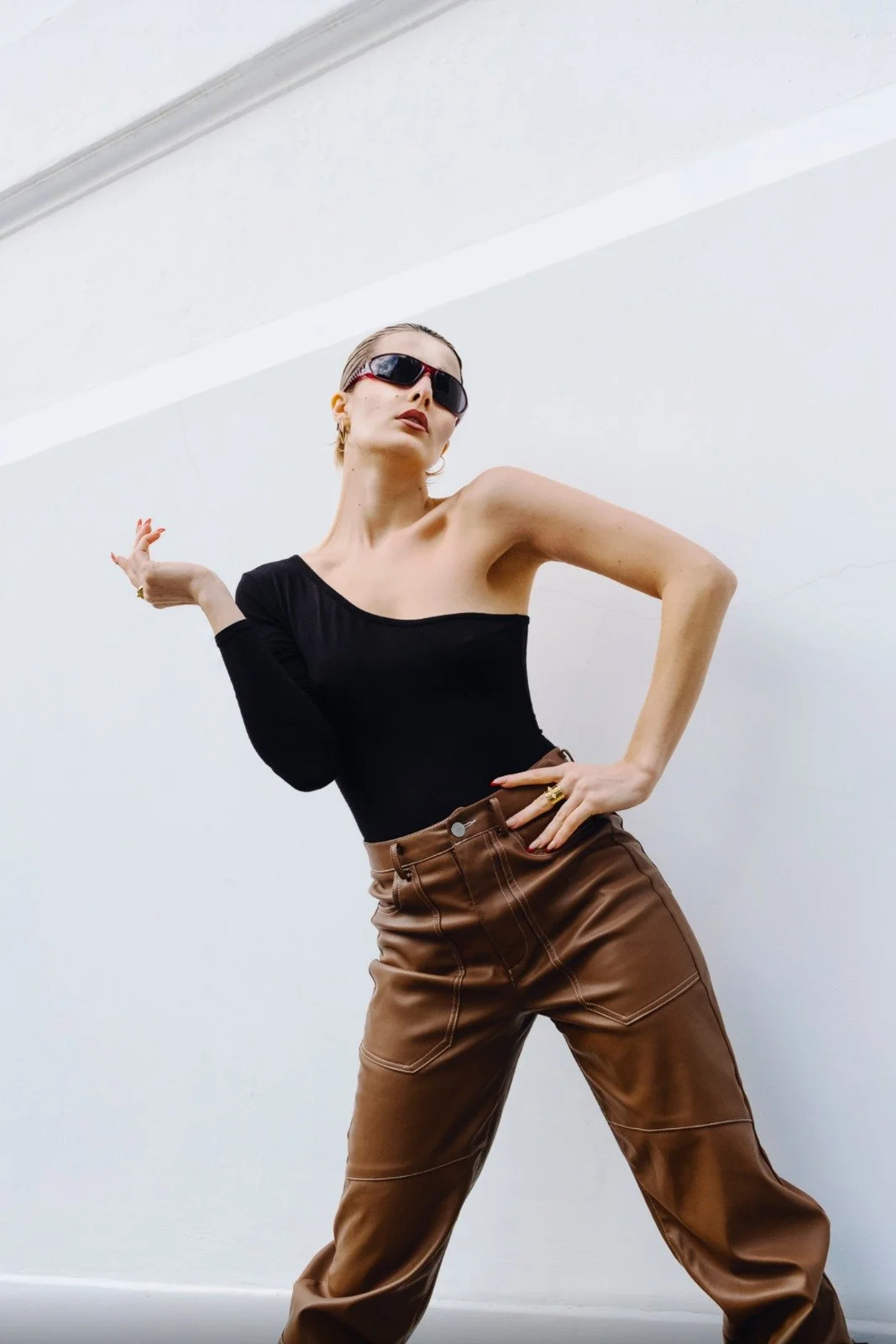 A woman with short blond hair wearing sunglasses, a black off-shoulder top, and brown leather pants striking a pose against a plain white wall.