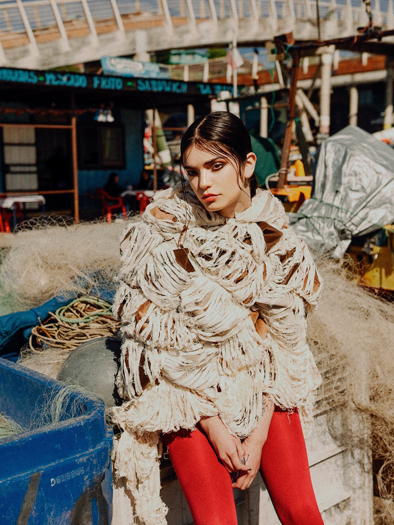 A woman with dark hair and makeup standing in front of a fishing area with nets, ropes, and boats. She is wearing a unique, shredded fabric top and red pants.