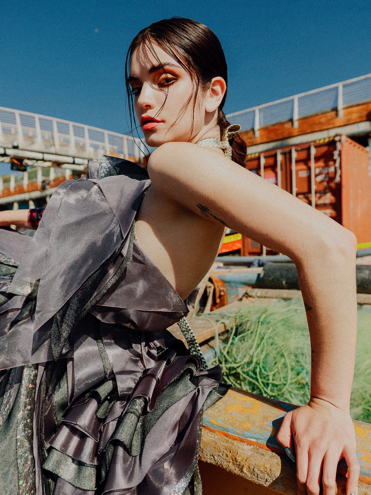 A woman with wet hair, wearing a gray and black dress, sitting outdoors on a sunny day with an industrial background of shipping containers and a bridge.