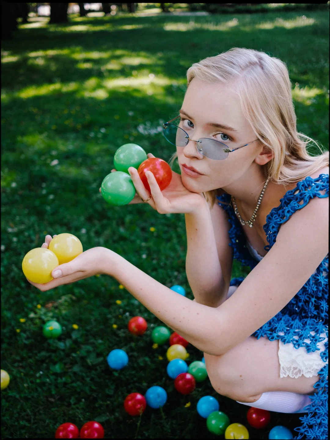 Young woman with blonde hair, wearing sunglasses and a blue top, sitting outdoors on grass with plastic colored balls in red, yellow, green, and blue.