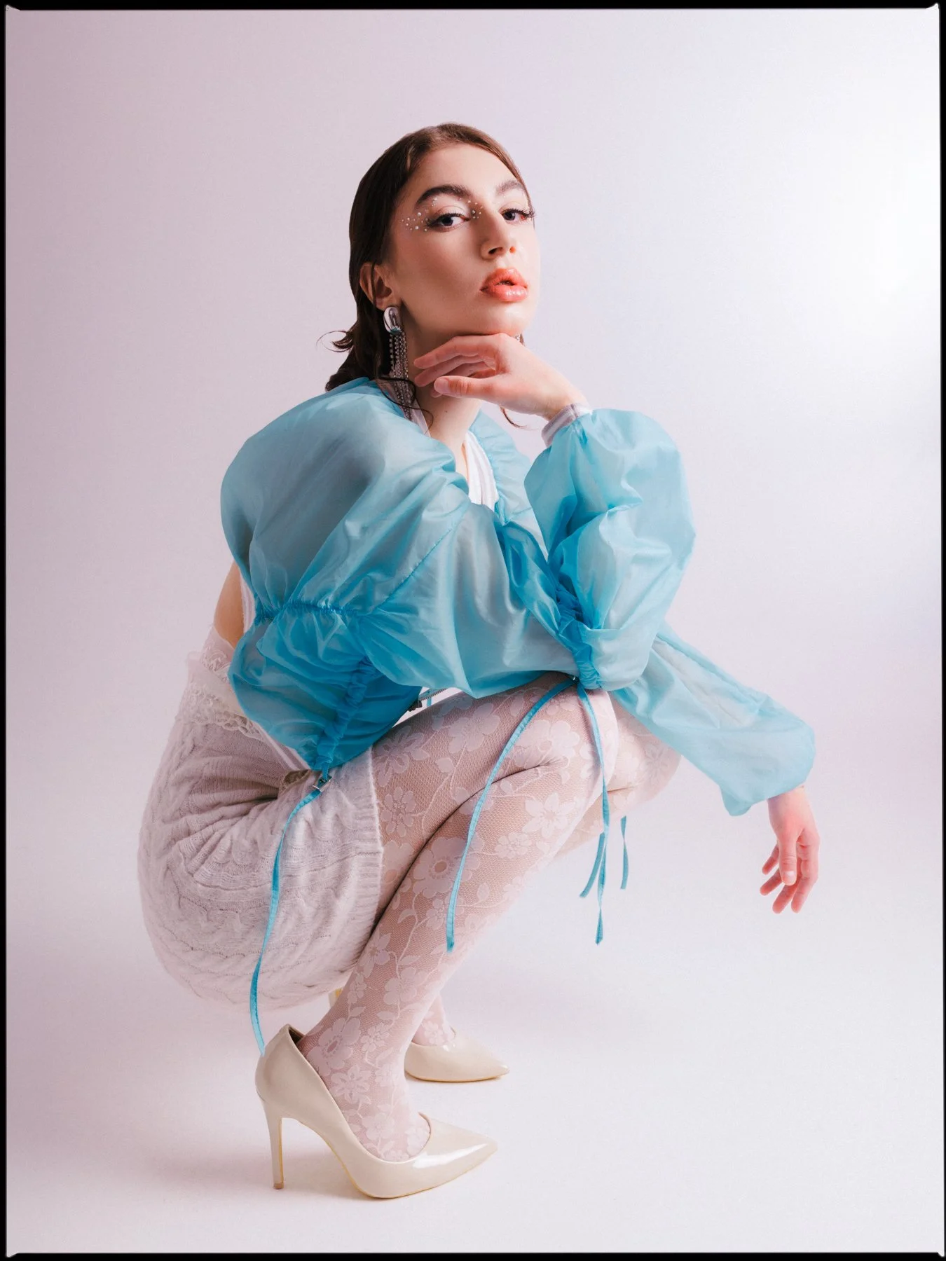 A woman with brown hair styled in loose waves, wearing makeup with rhinestones near her eyes. She is dressed in a light blue, oversized, puffy jacket, floral patterned tights, and high heels. She is posed in a crouching position against a plain white