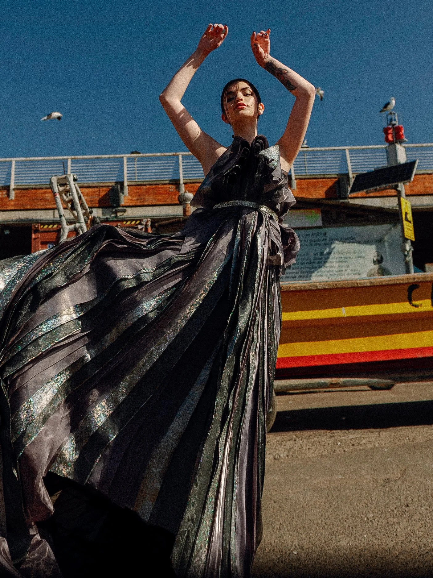 A woman with dark hair and makeup standing with her eyes closed wearing a shiny metallic and black dress, raising her arms. The background shows a blue sky, seagulls, and a building with a railing and a boat.