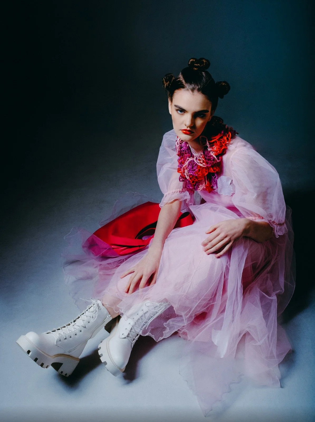 A young woman with styled hair in buns, wearing red lipstick, a pink tulle dress, and white ice skates, sitting on the floor with a dark background.