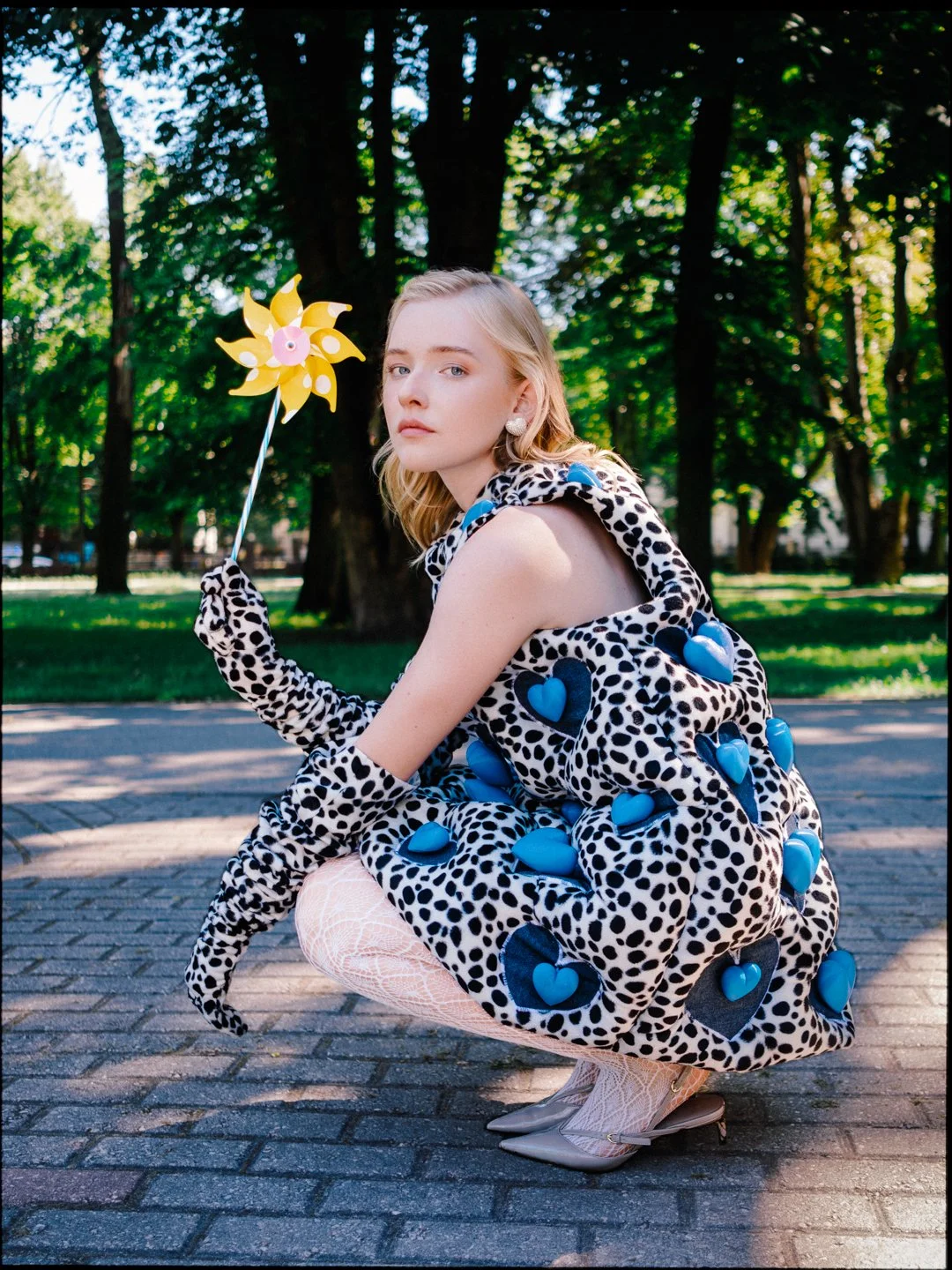 A young woman with blonde hair, wearing a black and white polka dot dress with blue heart accents, white fishnet stockings, and beige high heels, squatting outdoors on a paved pathway in a park with green trees in the background. She holds a yellow p
