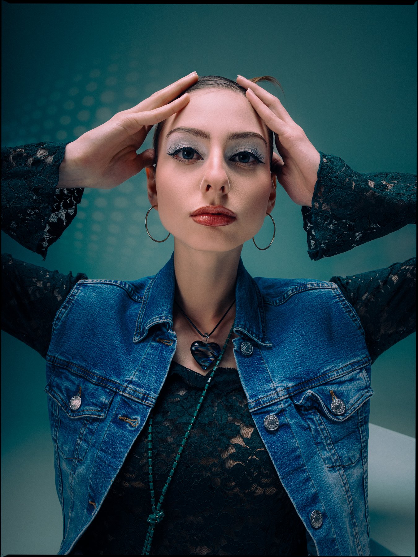 A woman with makeup, hoop earrings, and layered necklaces, wearing a denim vest over black lace clothing, holding her head with both hands, against a teal background.
