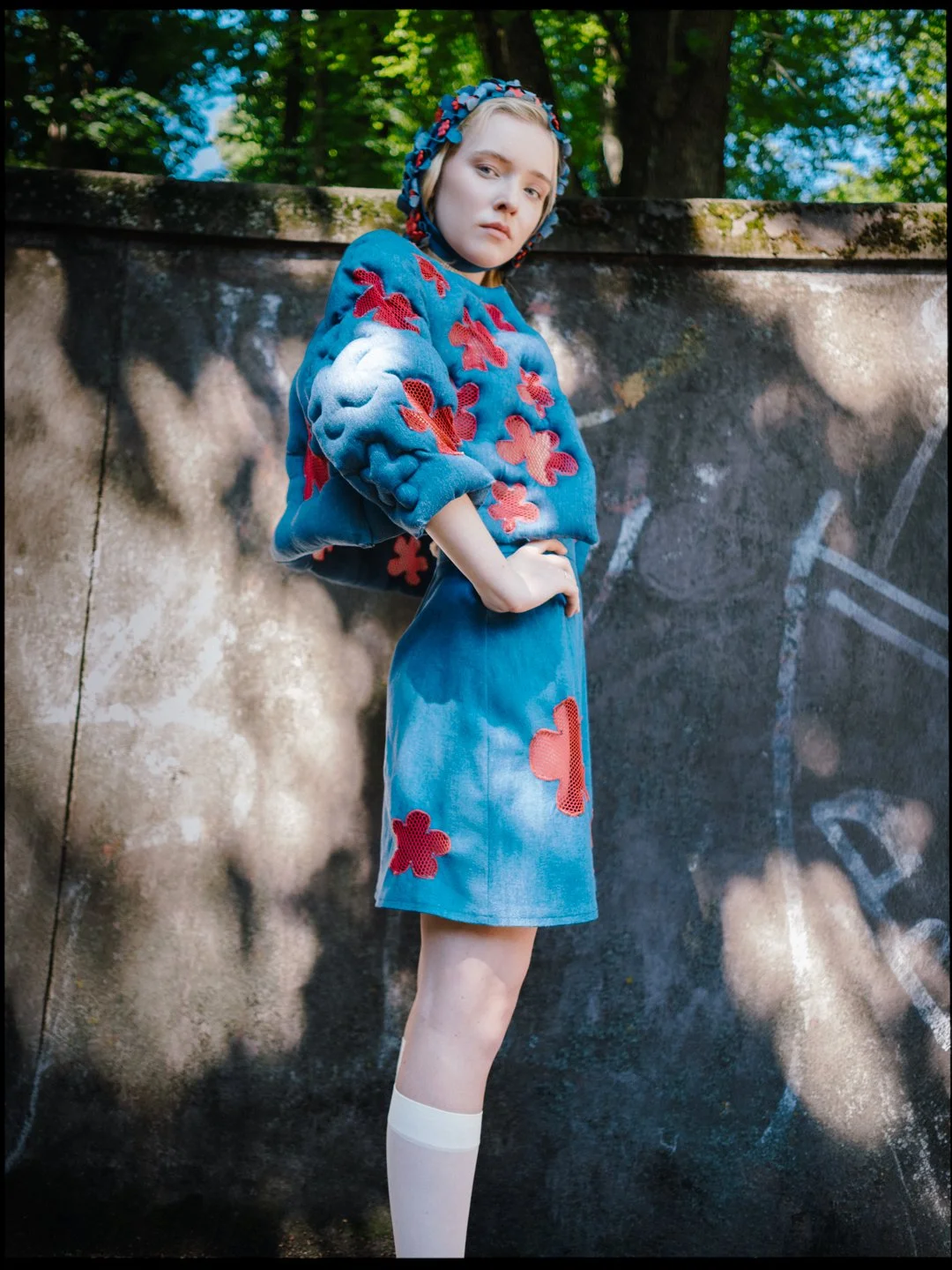 A young woman with blonde hair wearing a blue headscarf and a matching blue and red floral embroidered oversized sweater and skirt, standing outdoors against a wall with graffiti, surrounded by green trees and shadows.
