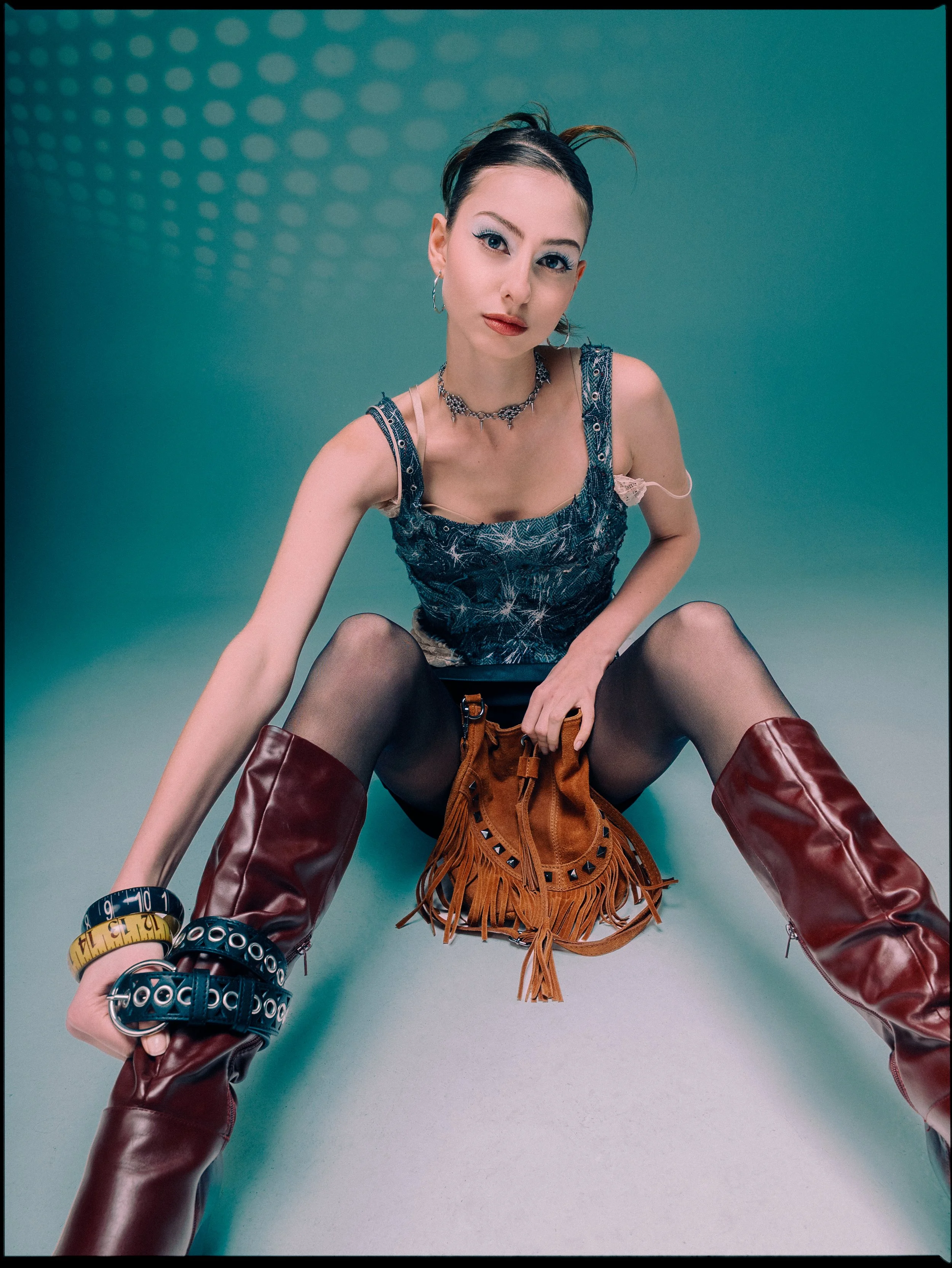 A woman with short dark hair styled with a side part, wearing makeup and silver jewelry, poses against a teal background with circular light patterns. She is dressed in a sleeveless top, fishnet stockings, and knee-high maroon boots, and has various 