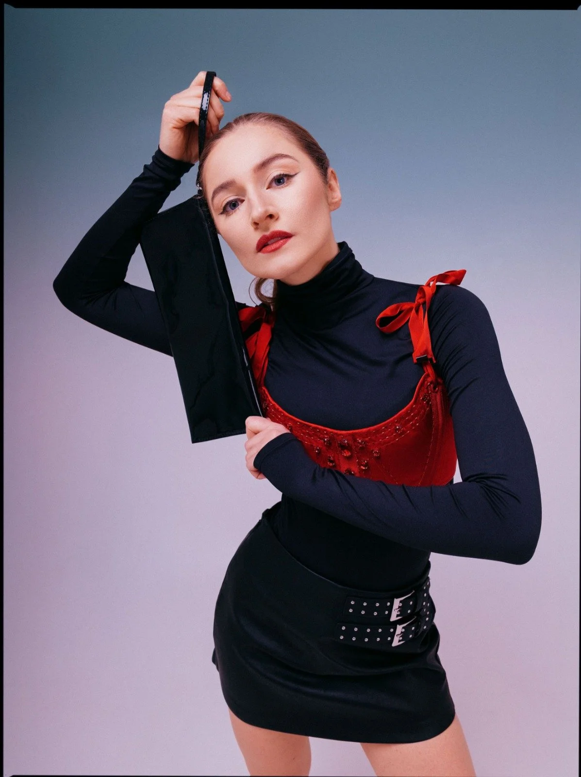 A woman with red lipstick and makeup, wearing a black turtleneck with red straps and a red embellished top, poses with one hand on her head holding a black shopping bag, against a plain gradient background.