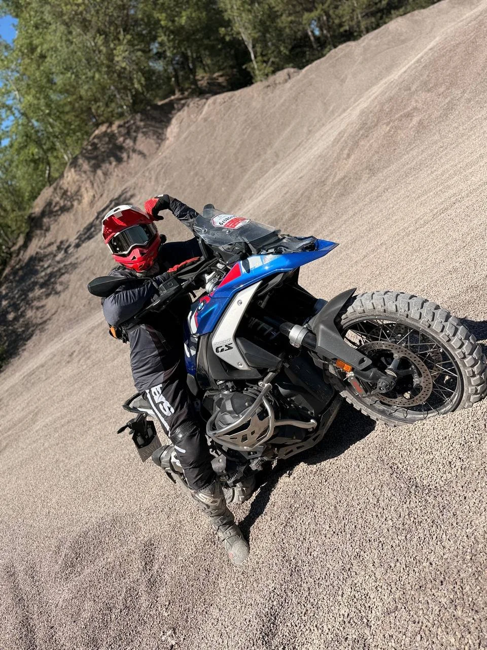 A motorcyclist in black riding gear and a red helmet is riding a blue and white dirt bike on a gravel trail surrounded by trees.