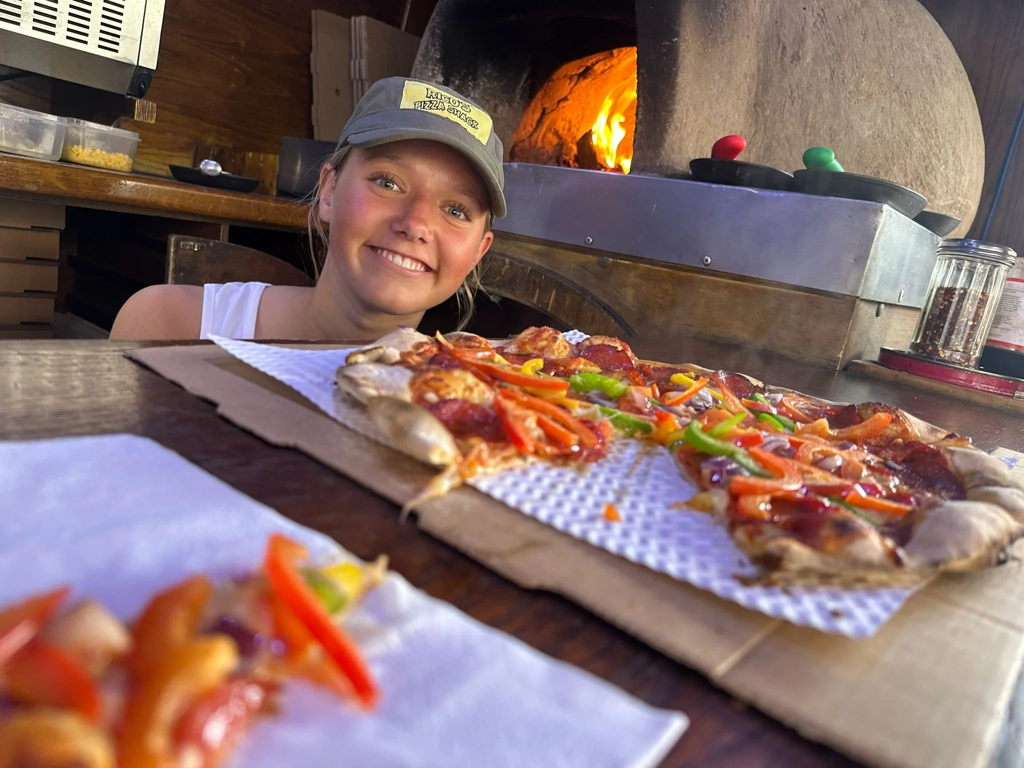 Smiling girl wearing a gray cap with a yellow patch, sitting in front of a pizza with colorful vegetables, inside a restaurant with a stone pizza oven with fire inside.