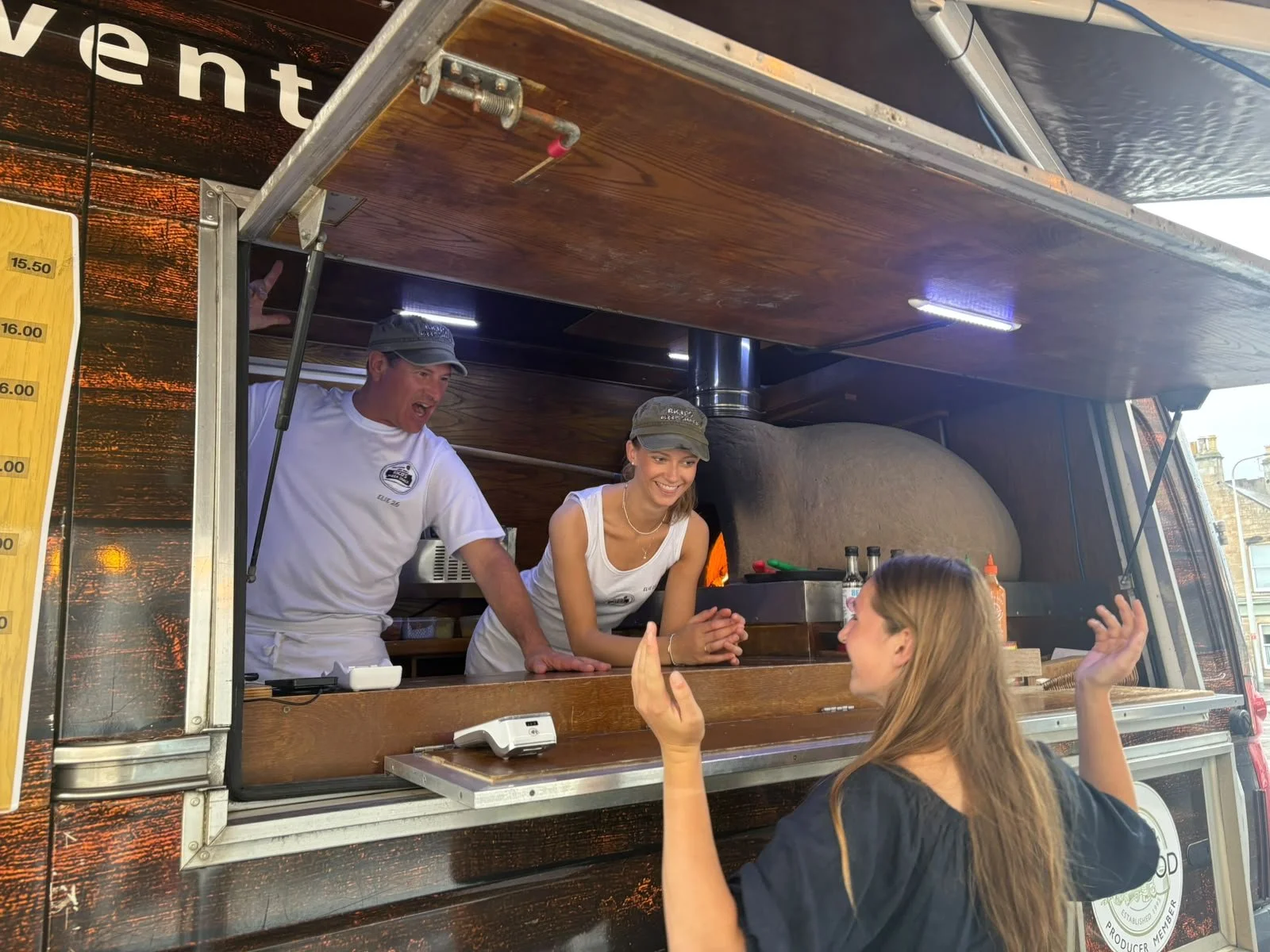 Two people are inside a food truck, smiling and talking to a customer. The person on the left is a man in a white shirt and a gray cap, smiling and leaning forward. The person in the middle is a woman in a gray cap and white tank top, smiling and with her hands clasped. The customer in front of them is a woman with long brown hair, gesturing with her hands. The food truck has a wood finish and various condiments are visible on the counter.