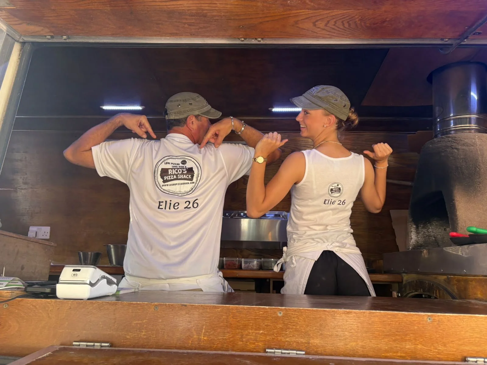 Two people, a man and a woman, wearing matching white shirts, flex their arm muscles and pose as if showing strength, inside a wood-paneled restaurant kitchen.