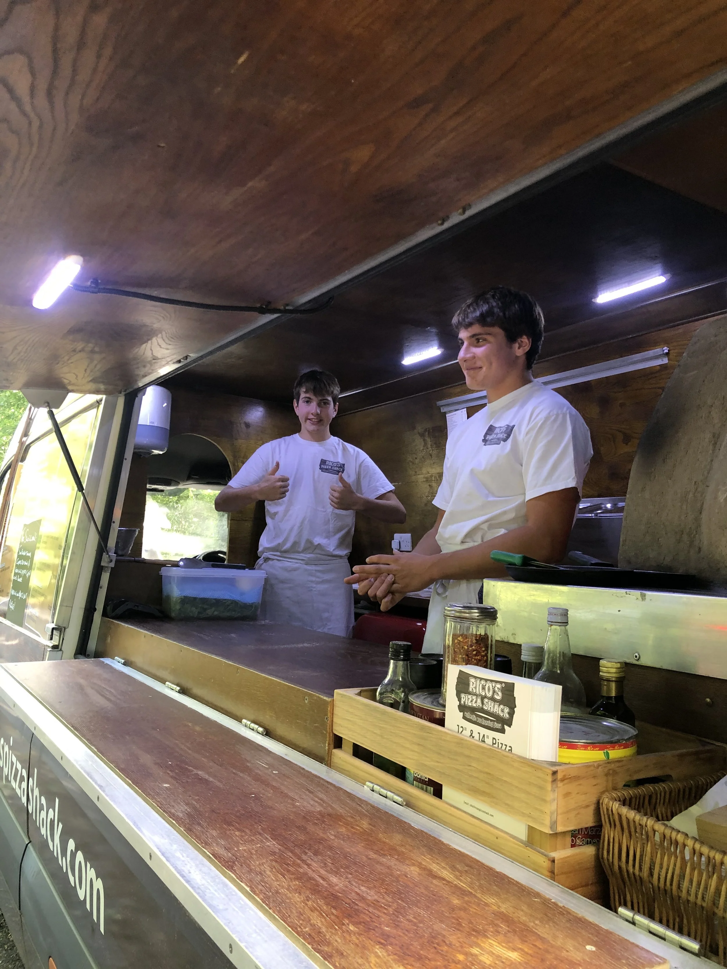 Two teenage boys in white t-shirts standing behind a food truck counter inside. One boy is giving a thumbs-up, the other is smiling with his hands clasped. The food truck is wooden, with bottles and a menu or sign visible in the foreground.