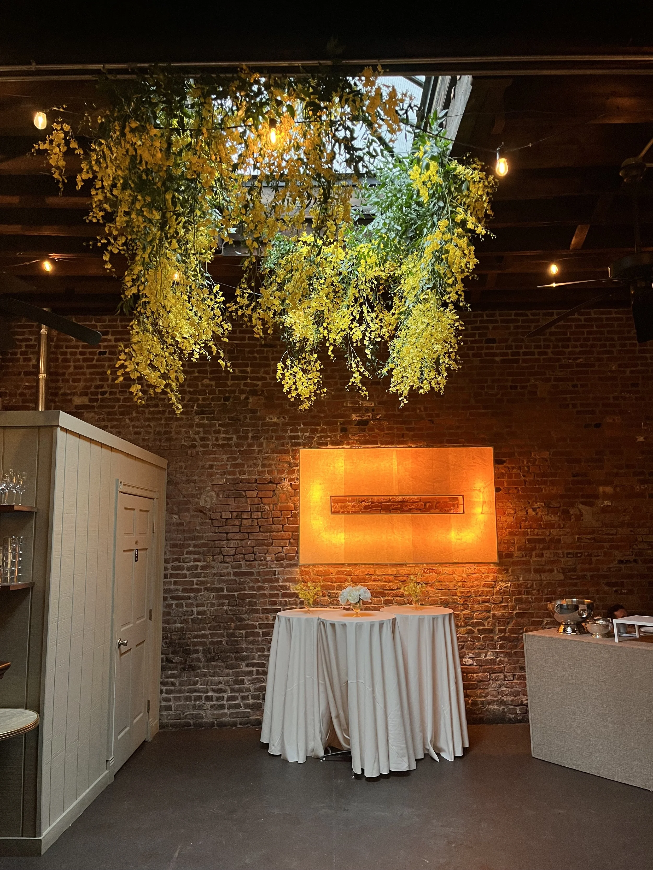 Interior of a decorated brick wall venue with a table covered with a white cloth, white flowers, and hanging yellow floral decoration from the ceiling. Warm lighting highlights the decor.