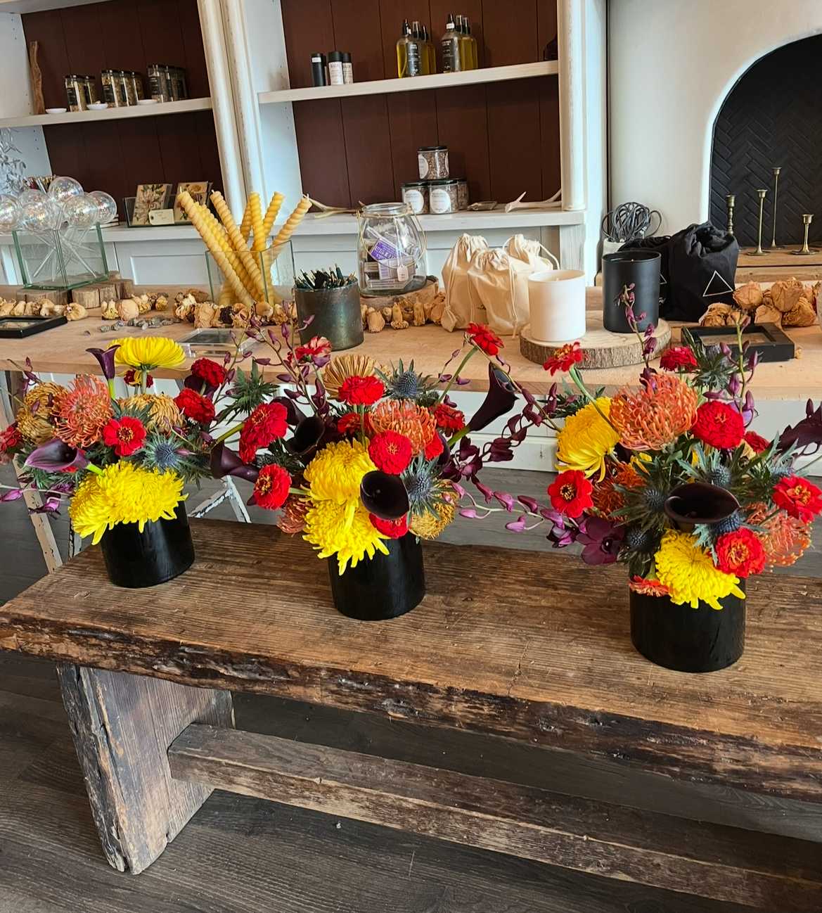 Three black vases filled with colorful flowers, including yellow, red, and dark purple blooms, arranged on a rustic wooden table in a cozy, well-decorated space with shelves and decorative items in the background.