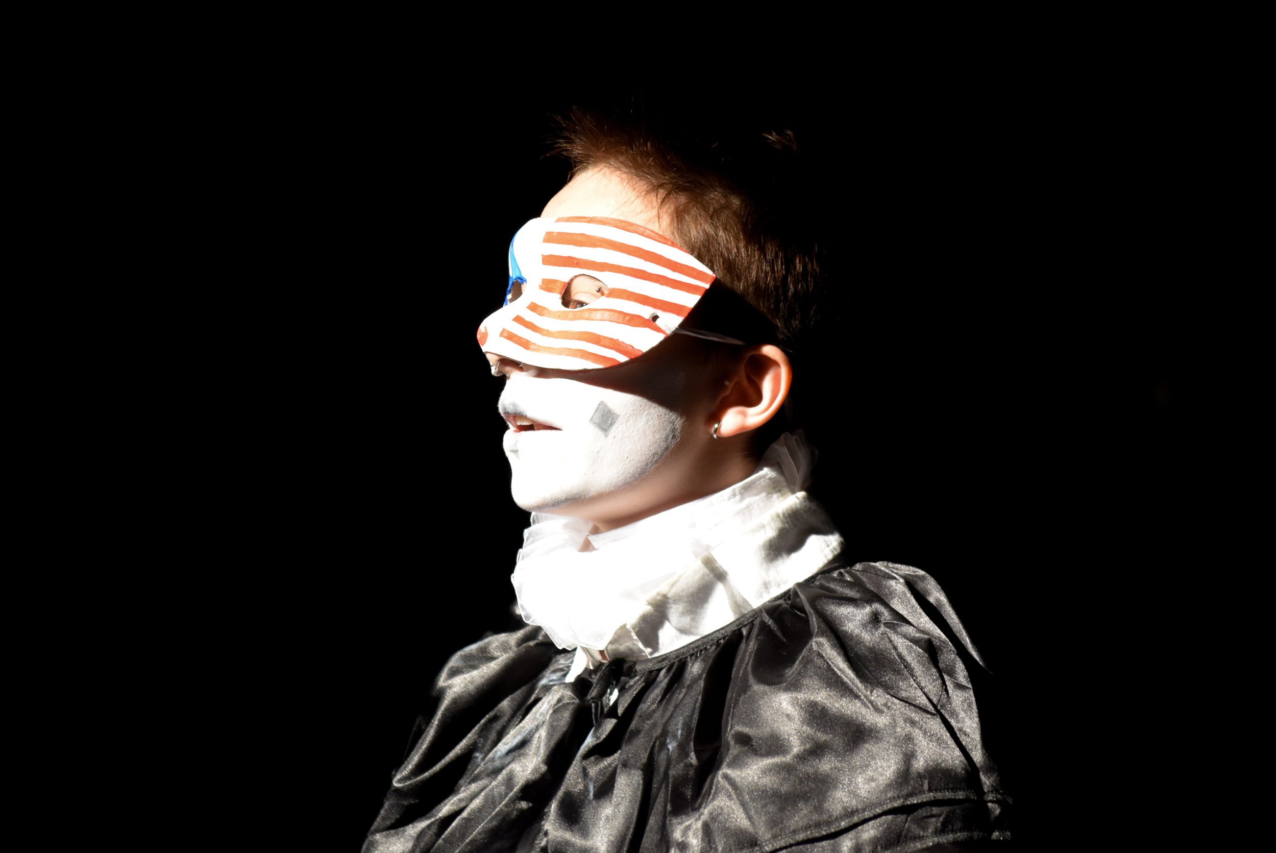 Person wearing a striped mask with red and white lines and blue details, with white face paint, dark curly hair, and a shiny black outfit, against a black background.