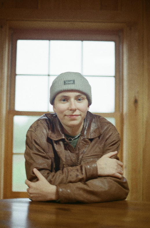 A young person with short hair wearing a gray beanie and a brown leather jacket, sitting at a wooden table inside a room with wooden walls and a window in the background.