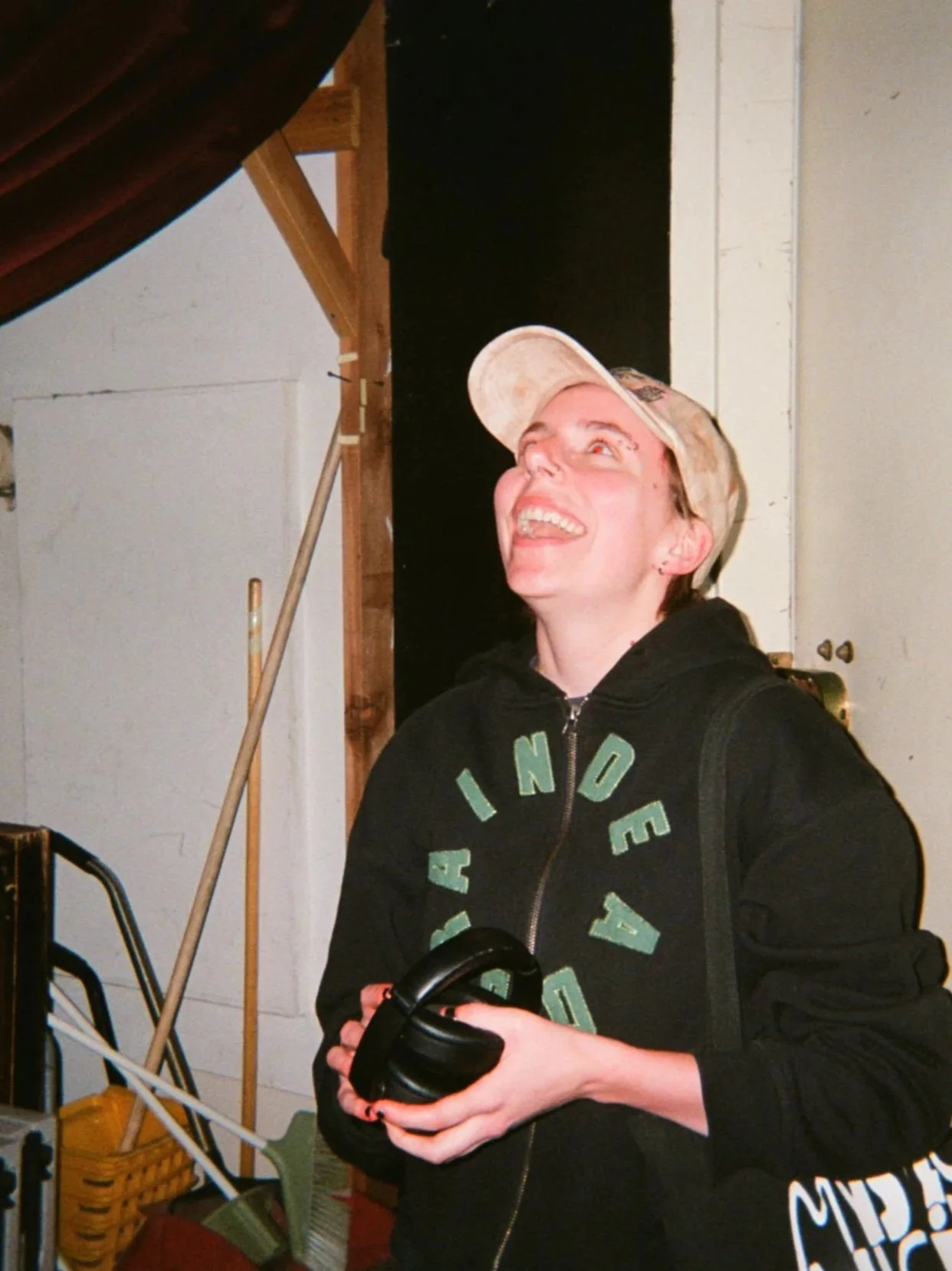 A young woman wearing a beige cap and black hoodie smiling and looking up while holding black headphones in her hands in an indoor setting with a wall, ladder, and miscellaneous items in the background.