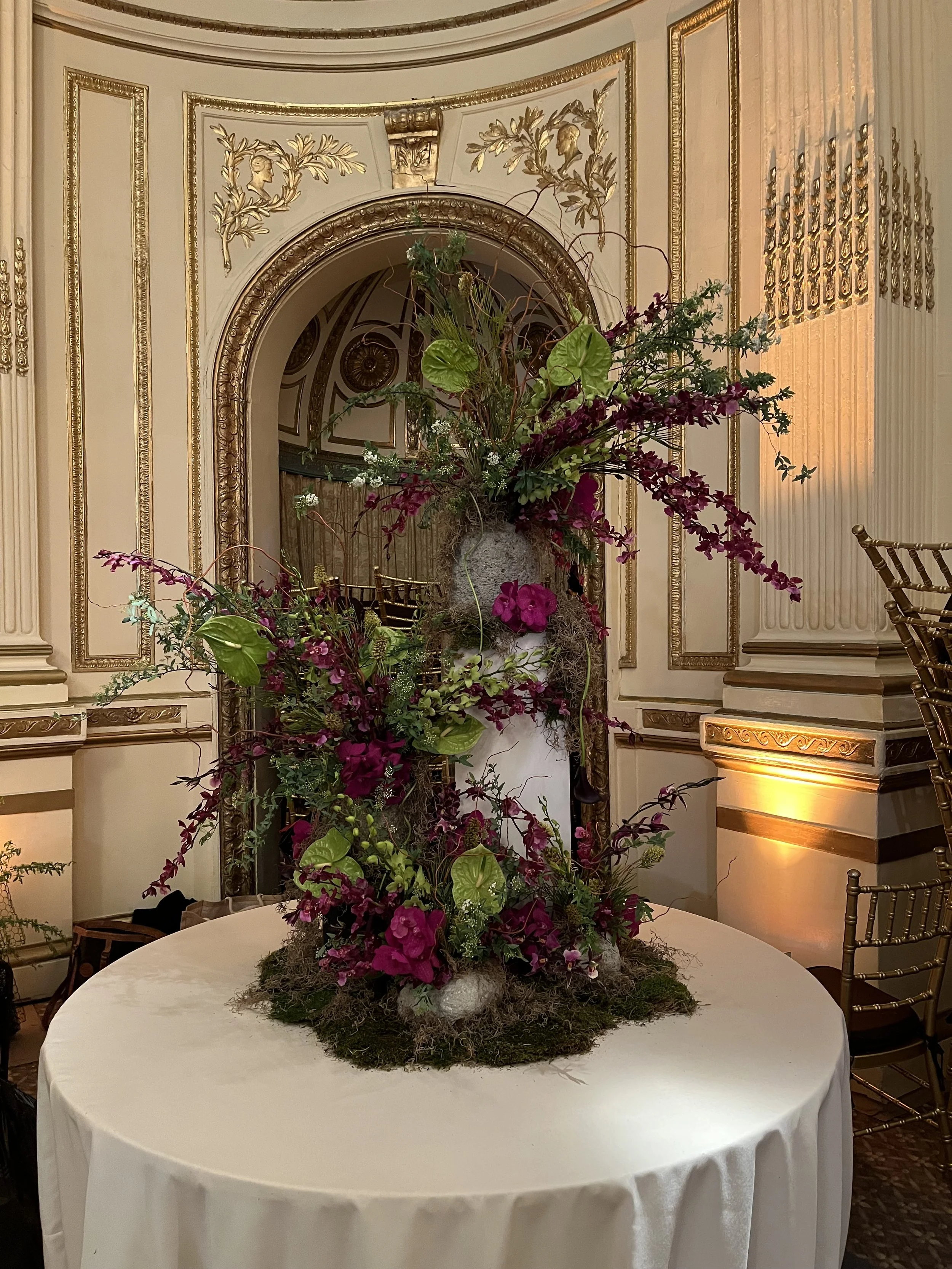 A large floral centerpiece on a round table with a white tablecloth, set against ornate, gold-trimmed cream walls with decorative moldings and a mirror in the background.