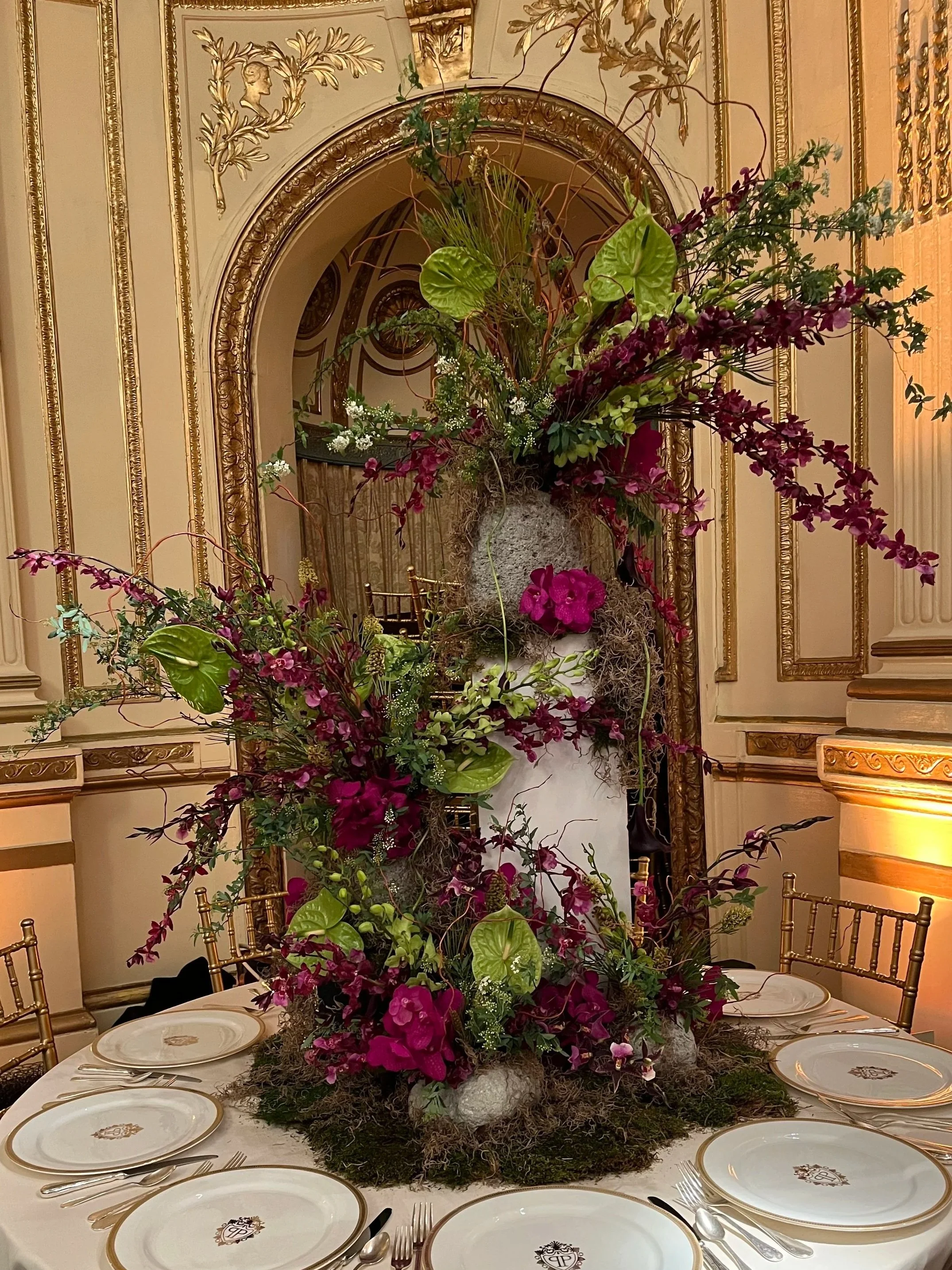 A formal banquet table in an ornate room with a large floral centerpiece. The table is set with white plates featuring a gold rim and a monogrammed crest, along with silverware. In the background, elaborate gold and cream paneling adorns the walls.