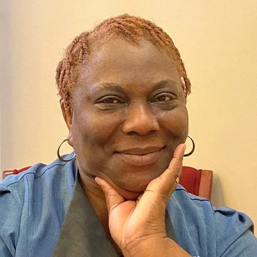 A woman with reddish-brown hair styled in twists, wearing a blue shirt, smiling and resting her chin on her hand.