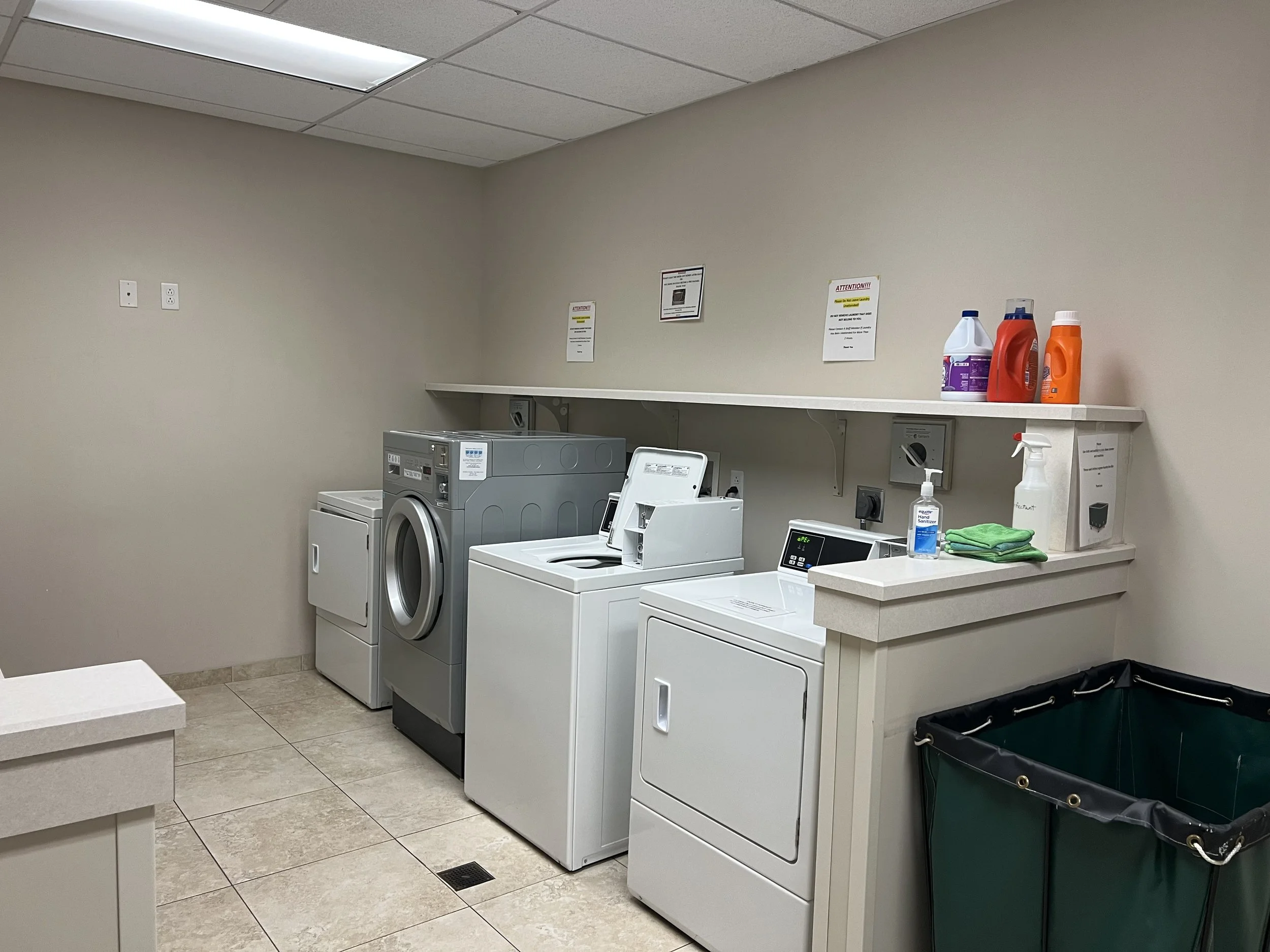 Laundry room with washing machines, detergent bottles, and cleaning supplies. There are several signs on the wall and a plastic laundry hamper on the floor.