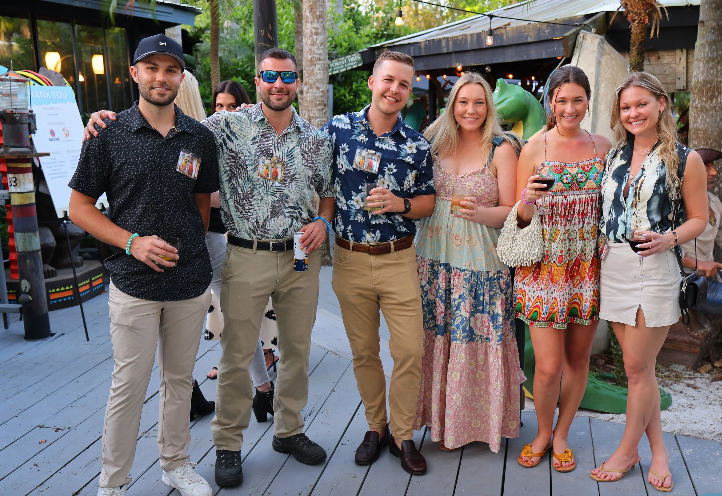 Seven smiling young adults holding drinks and gathering outdoors under string lights at a Gabriel House summer event.