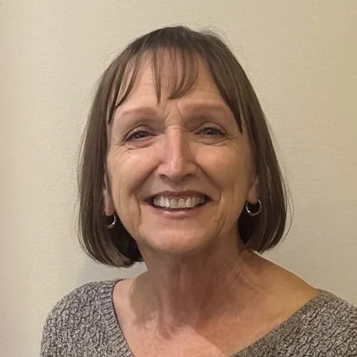 A middle-aged woman with short brown hair and bangs, smiling, wearing a gray sweater and small hoop earrings, standing against a plain beige wall.