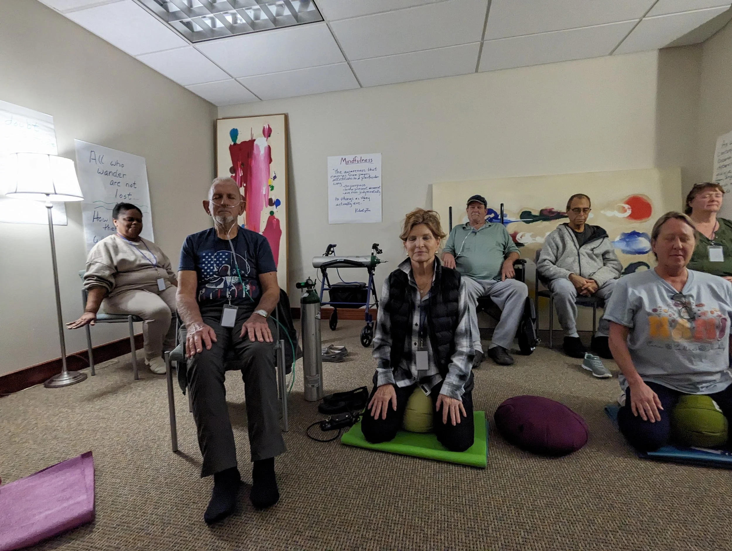 A group of people sitting on chairs and on the floor in a room with beige walls and carpet, participating in a yoga or meditation session. Some individuals are seated on yoga mats with meditation cushions. There are posters on the walls with inspirational quotes about mindfulness and wandering.