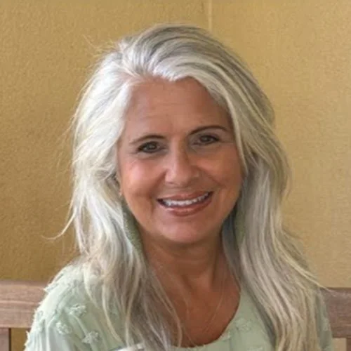 A smiling woman with long gray hair sitting indoors against a yellow wall.