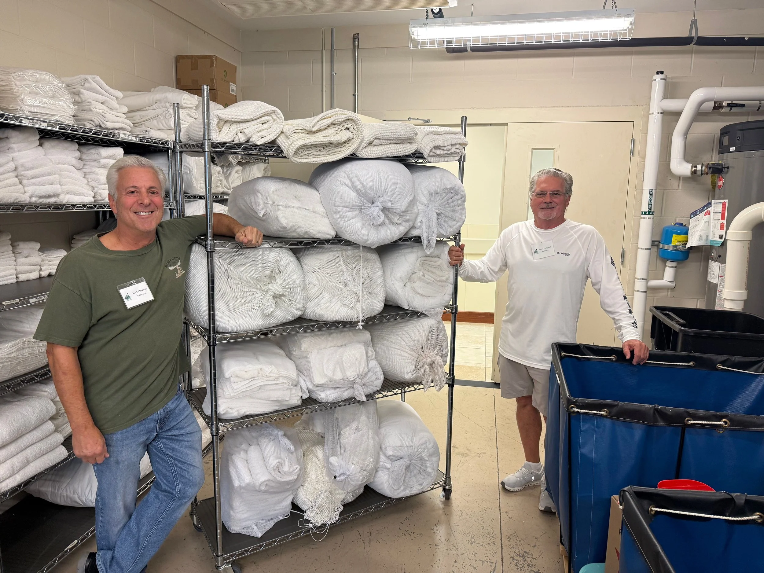 Two smiling men standing in a linen storage room filled with neatly folded white towels.