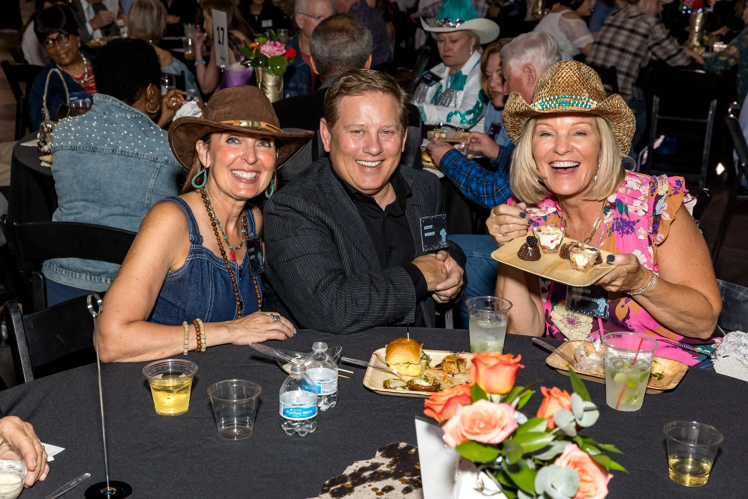 A group of three people sitting at a dinner table, smiling at the camera. Two women are wearing cowboy hats, one with a floral dress and the other with a denim dress. The man in the center is dressed in a dark striped suit. They are at a social event with plates of food, drinks, and decorative flowers on the table.