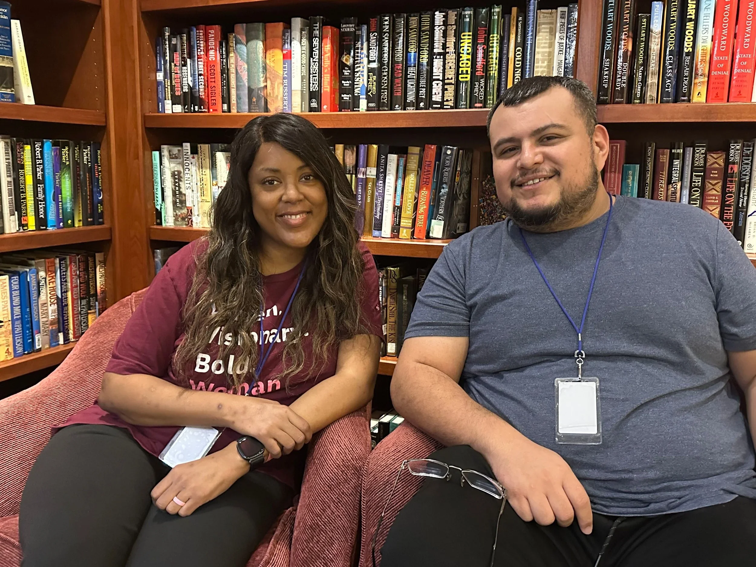A woman with long dark hair and a man with short dark hair and a beard sitting in front of a bookshelf filled with books. They are both smiling, and the woman is wearing a maroon shirt while the man is wearing a blue shirt.